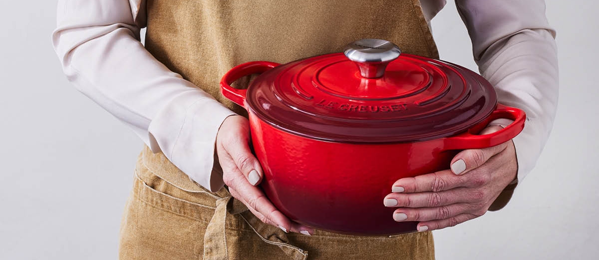 Person i forkle holder en rød Le Creuset-gryte med lokk.