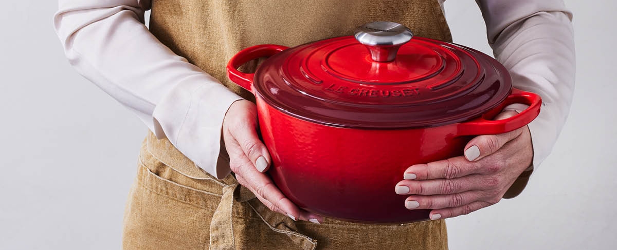 Person i forkle holder en rød Le Creuset-gryte med lokk.