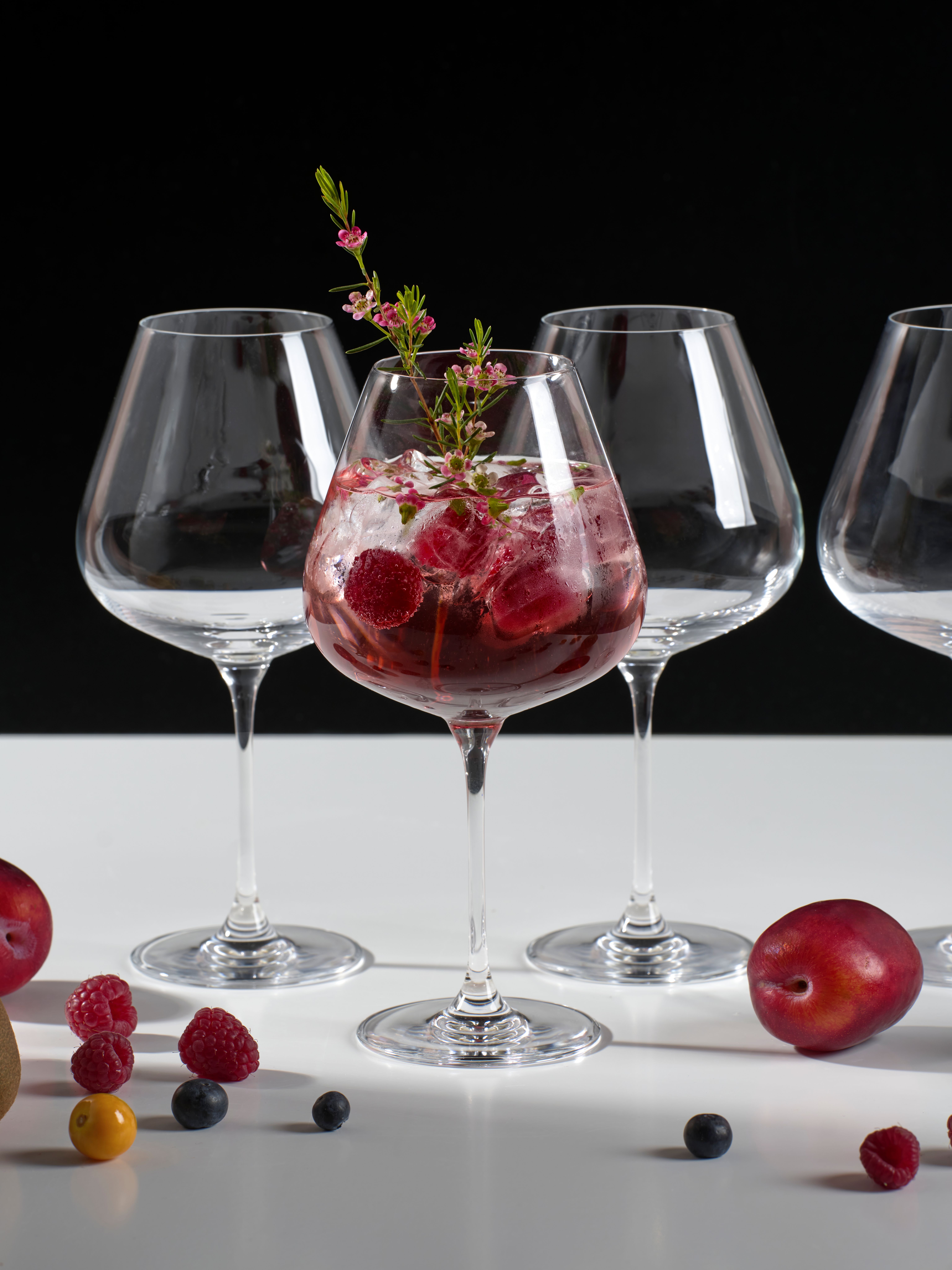 Elegant vinglas med rosa drink, is, hallon och blomsterdekoration mot svart bakgrund.