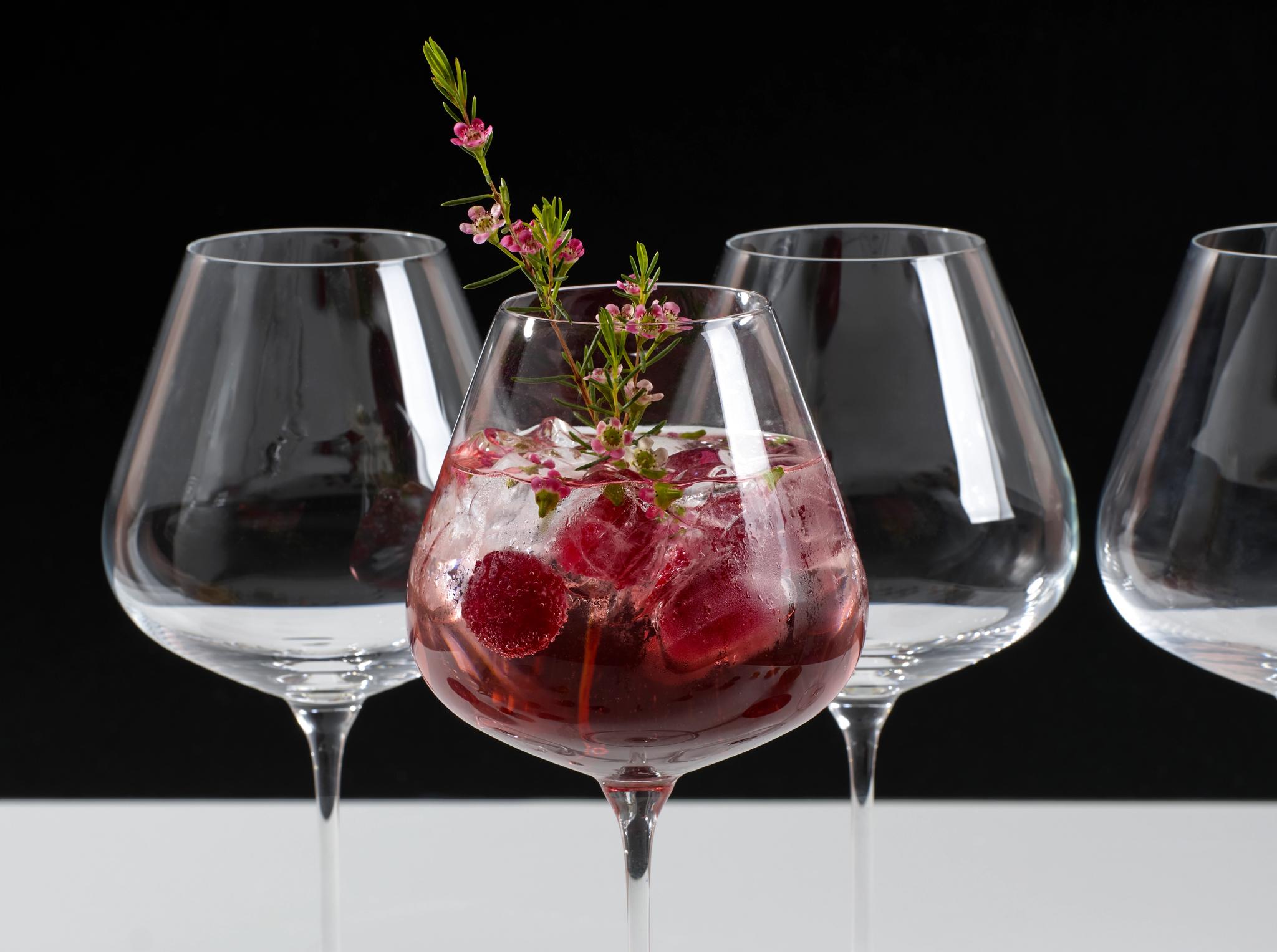 Elegant vinglas med rosa drink, is, hallon och blomsterdekoration mot svart bakgrund.