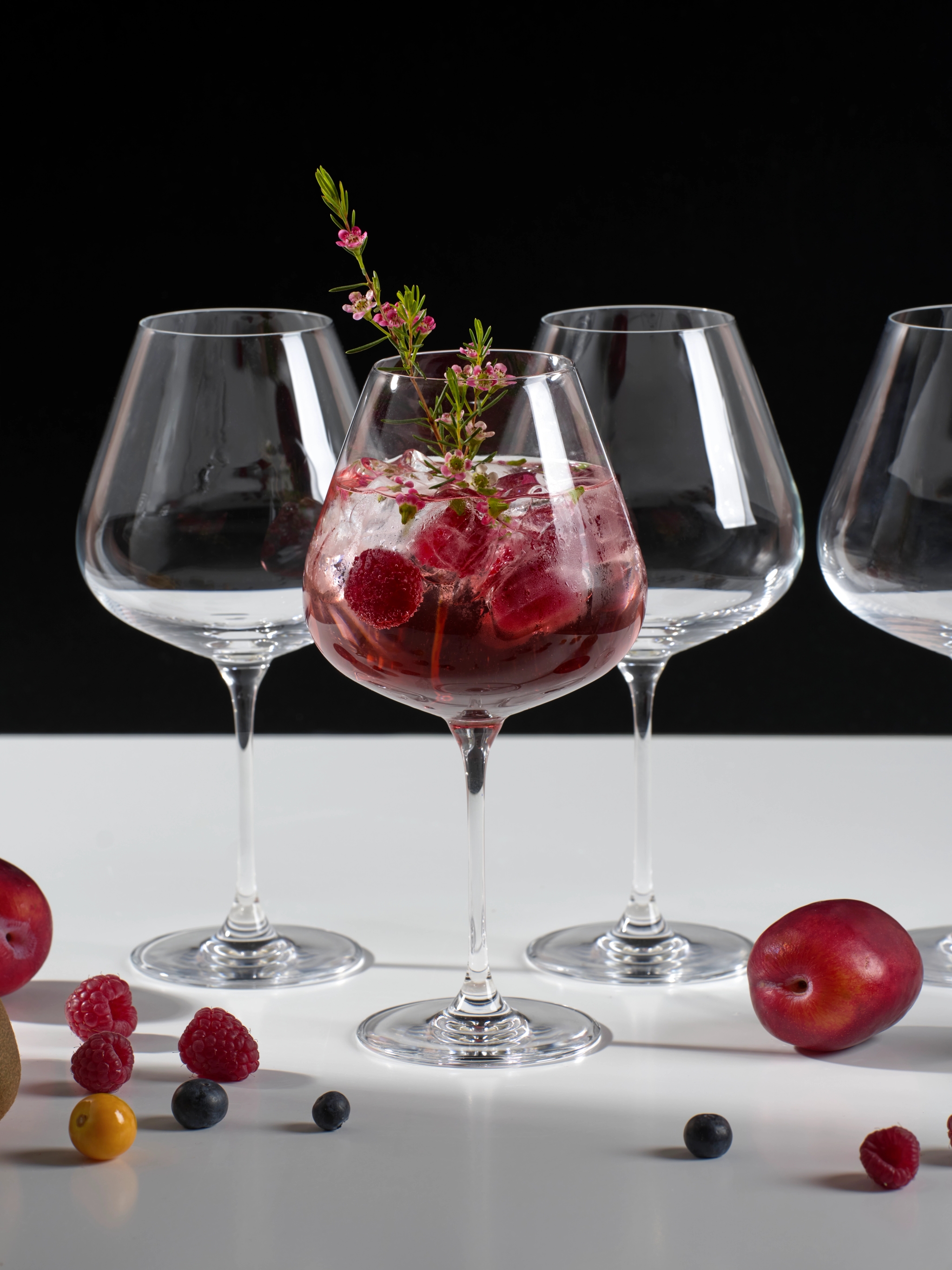 Elegant cocktail med is, hallon och rosa blommor i ett vinglas, omgiven av tomma glas och färska bär.