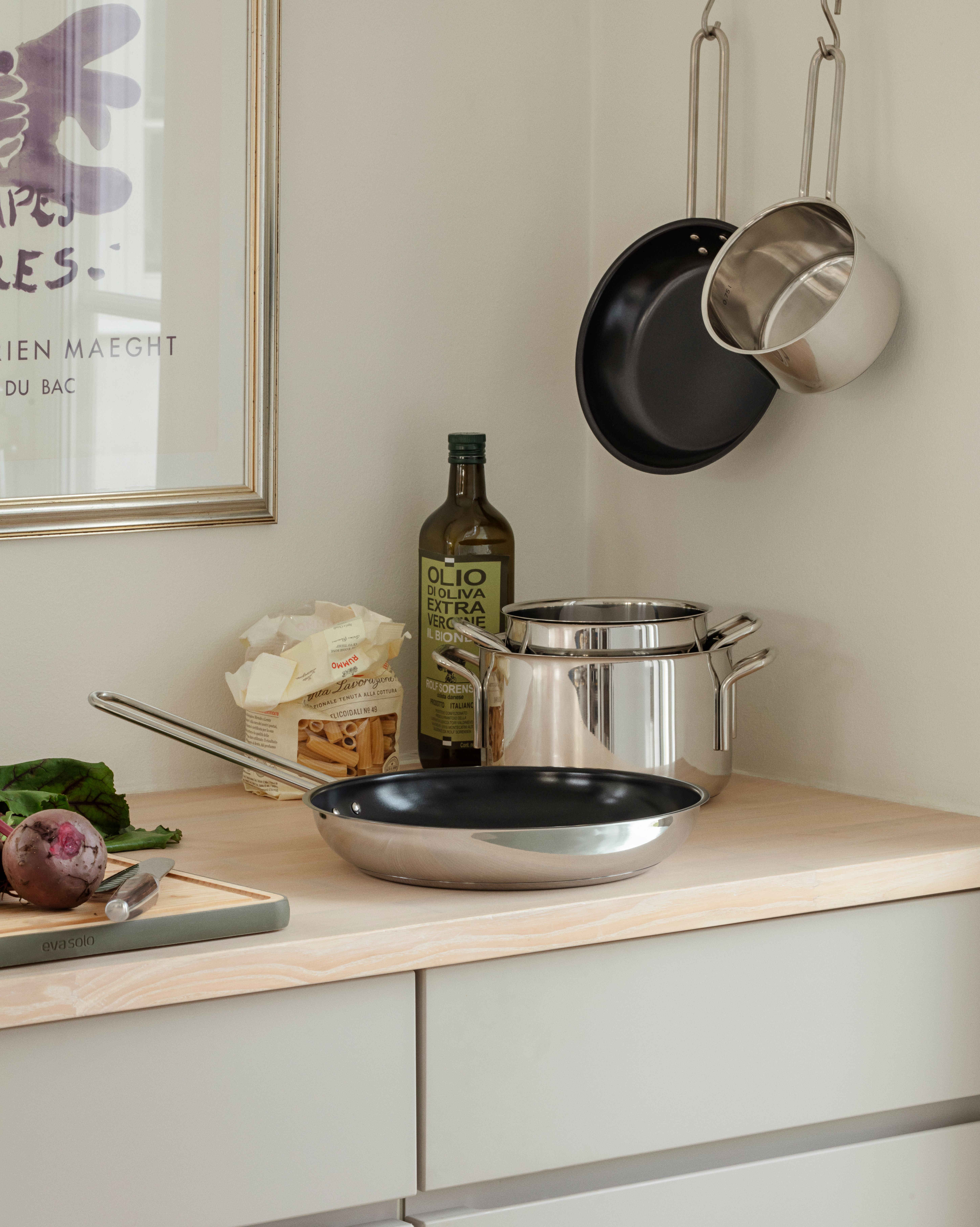 Keittiön tiskillä ruostumattomasta teräksestä valmistettuja keittovälineitä, oliiviöljyä, pastaa ja punajuuri leikkuulaudalla kehystetyn taideteoksen vieressä.