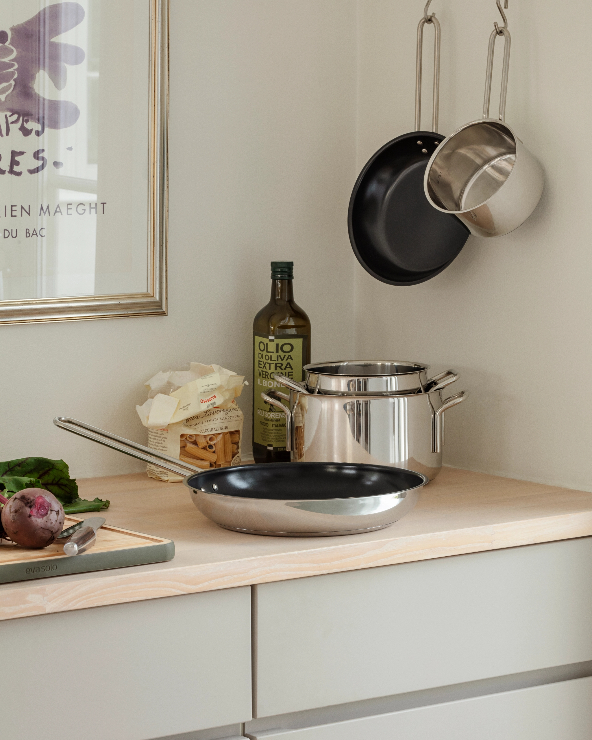 Keittiön tiskillä ruostumattomasta teräksestä valmistettuja keittovälineitä, oliiviöljyä, pastaa ja punajuuri leikkuulaudalla kehystetyn taideteoksen vieressä.