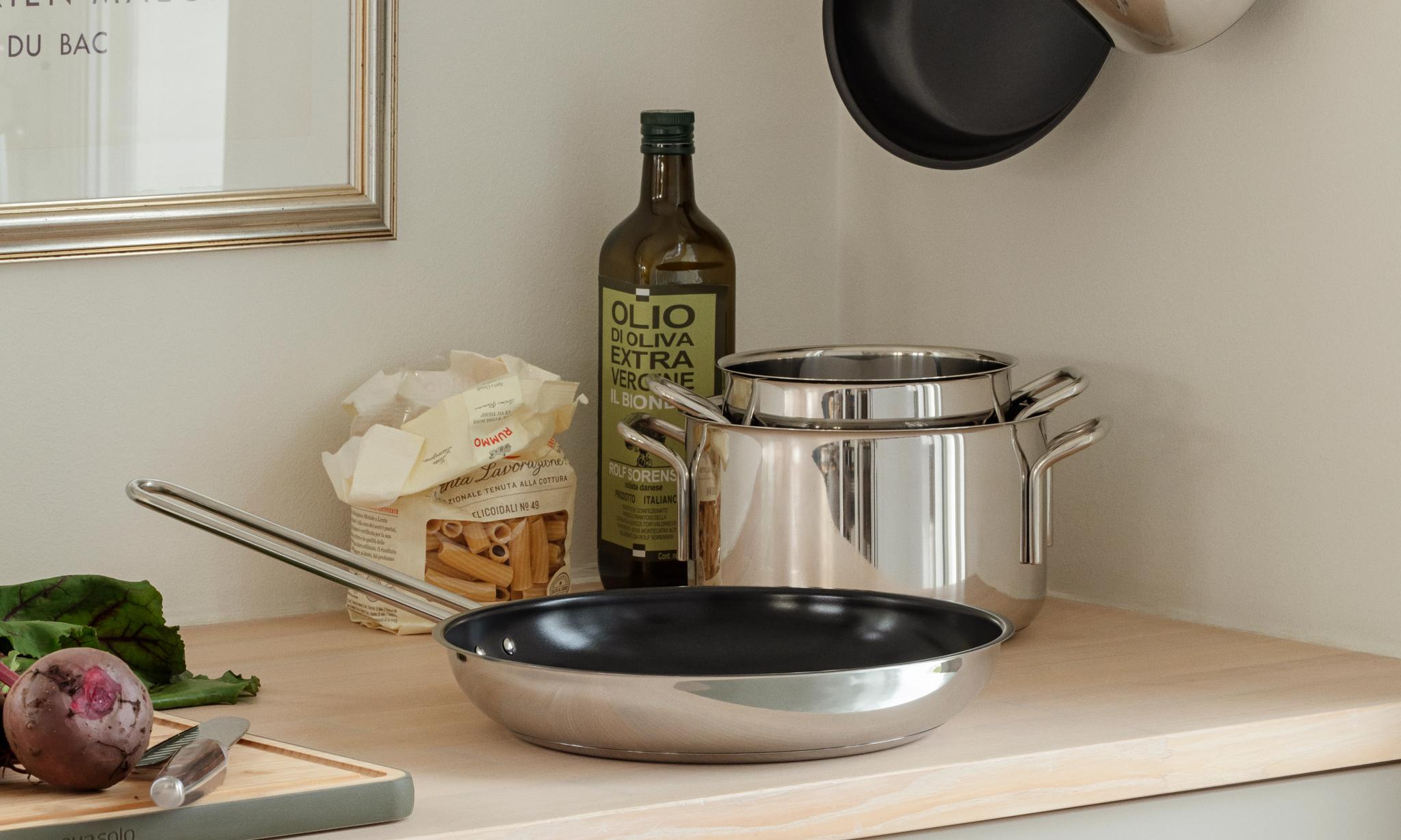 Keittiön tiskillä ruostumattomasta teräksestä valmistettuja keittovälineitä, oliiviöljyä, pastaa ja punajuuri leikkuulaudalla kehystetyn taideteoksen vieressä.