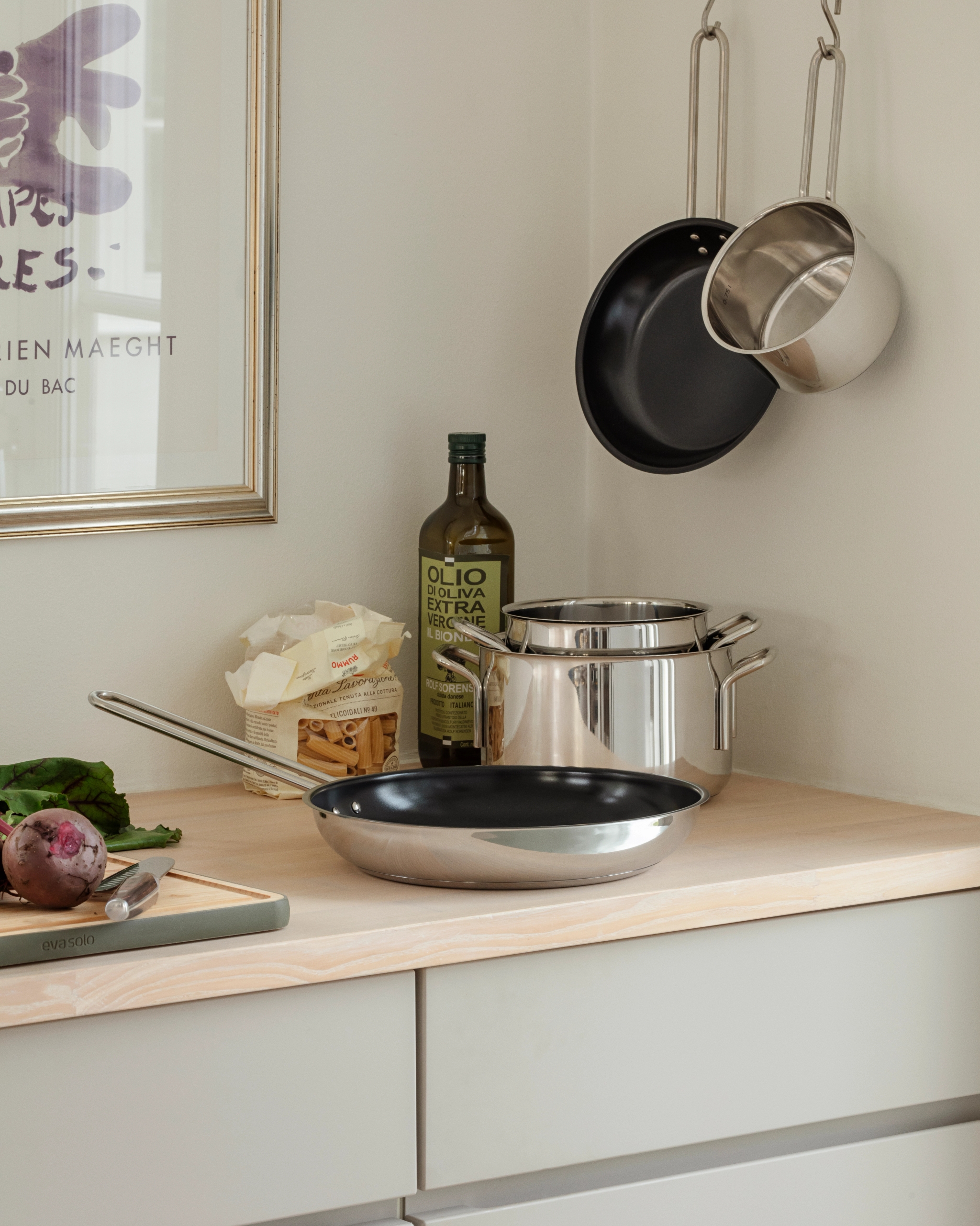 Keittiön tiskillä ruostumattomasta teräksestä valmistettuja keittovälineitä, oliiviöljyä, pastaa ja punajuuri leikkuulaudalla kehystetyn taideteoksen vieressä.