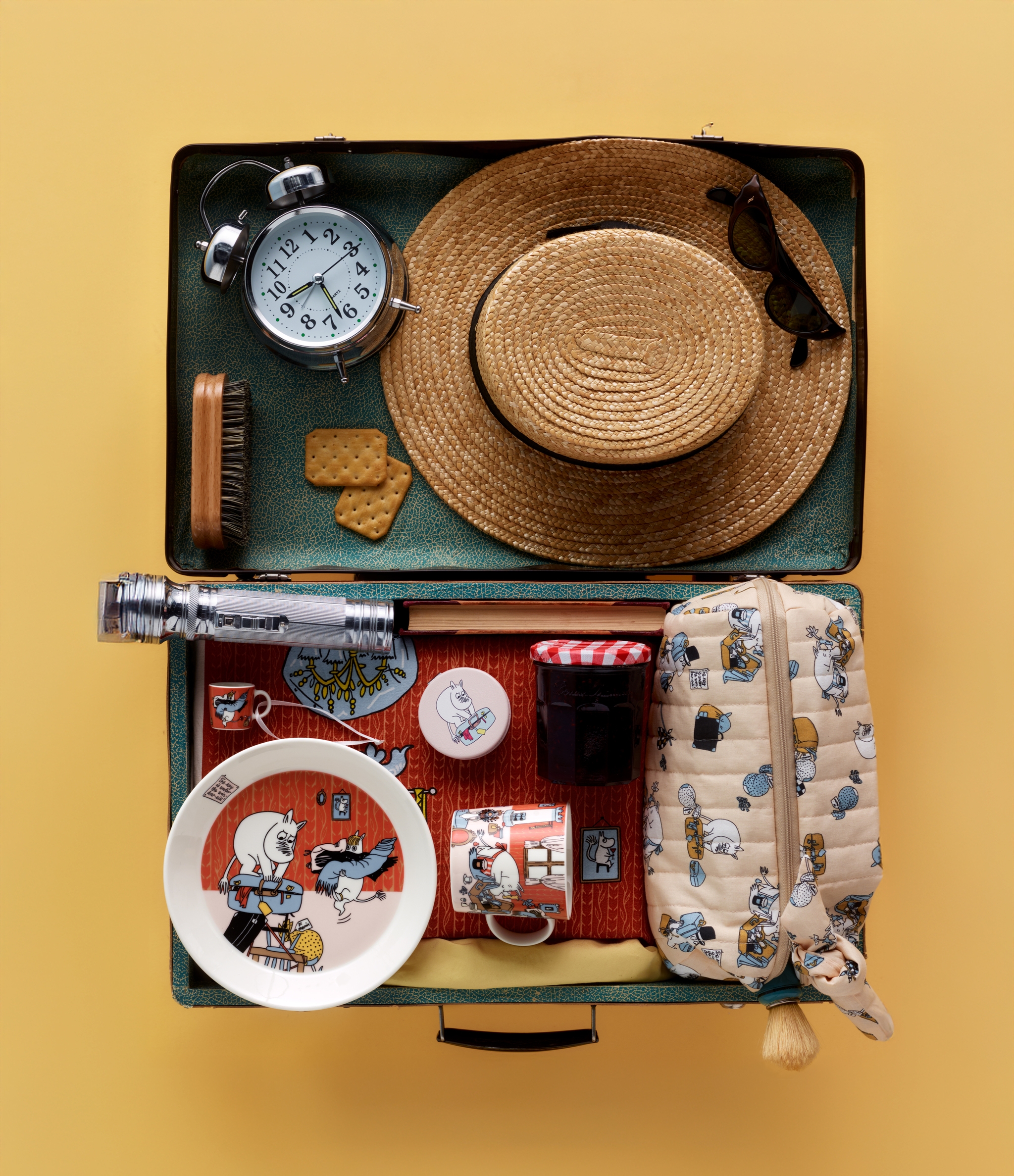 En öppen vintageresväska fullpackad med rese- och picknickartiklar med Mumin-tema, inklusive en stråhatt, väckarklocka, porslin och sylt.