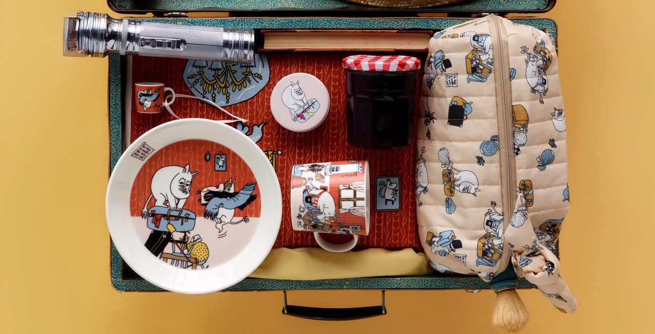 En öppen resväska fullpackad med resesaker med Mumintema, en stråhatt, väckarklocka och kex.