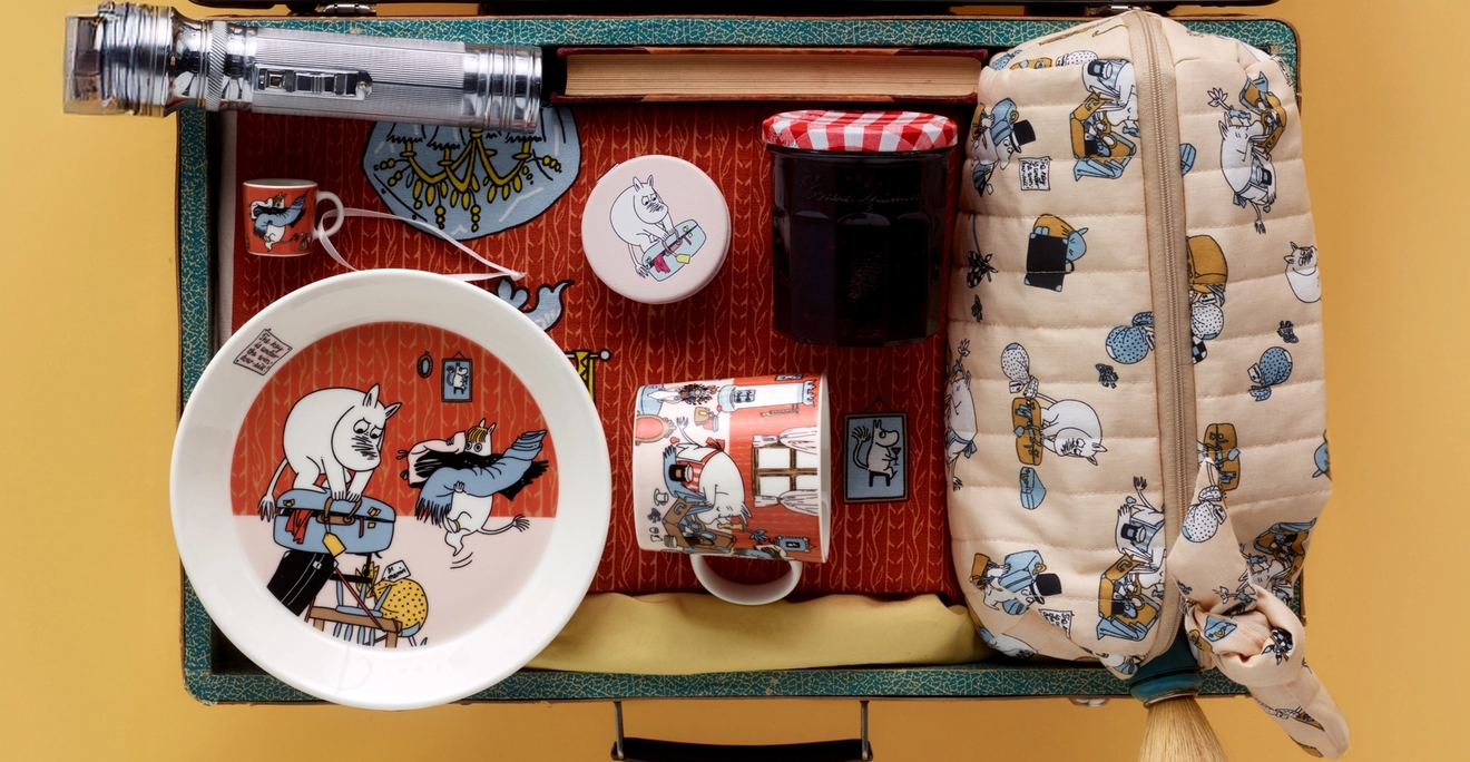 En öppen resväska fullpackad med resesaker med Mumintema, en stråhatt, väckarklocka och kex.