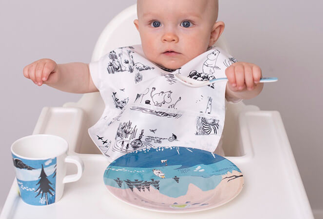 Rätt Start bebis sitter i barnstol med haklapp, tallrik, mugg och sked 