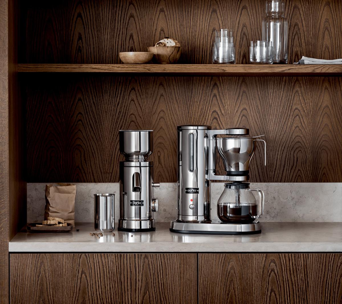 Moderne kaffemaskin og kaffekvern i rustfritt stål på kjøkkenbenk med trehyller i bakgrunnen.