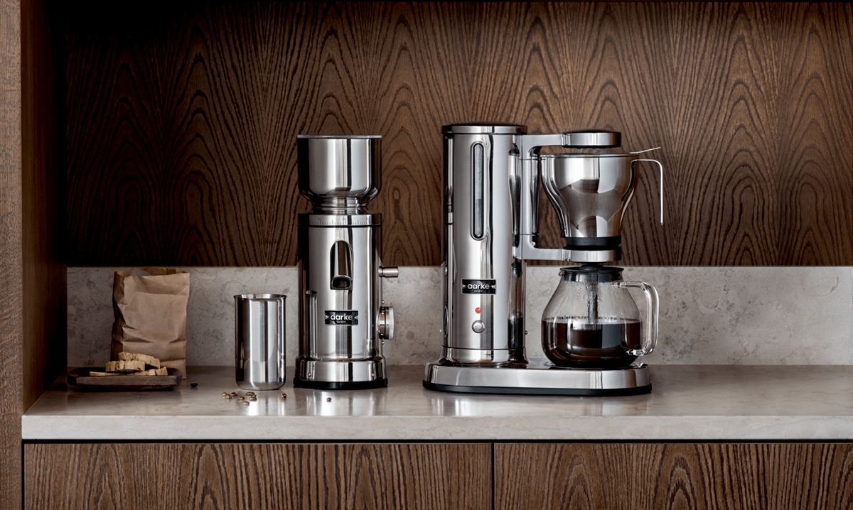 En modern kaffekvarn och kaffebryggare i rostfritt stål på en köksbänk med mörk träpanel.