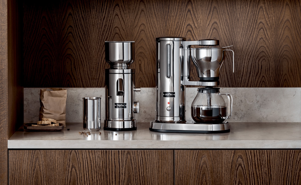 Ruostumattomasta teräksestä valmistettu kahvimylly ja kahvinkeitin tiskillä, taustalla puiset hyllyt, joilla on keittiövälineitä.