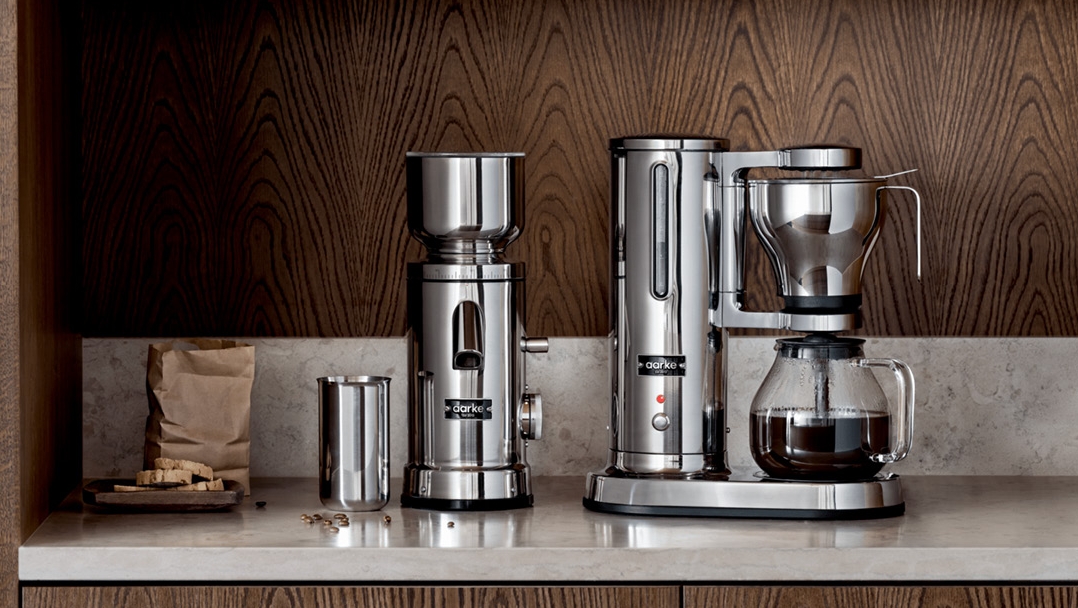 En modern kaffekvarn och kaffebryggare i rostfritt stål på en köksbänk med mörk träpanel.