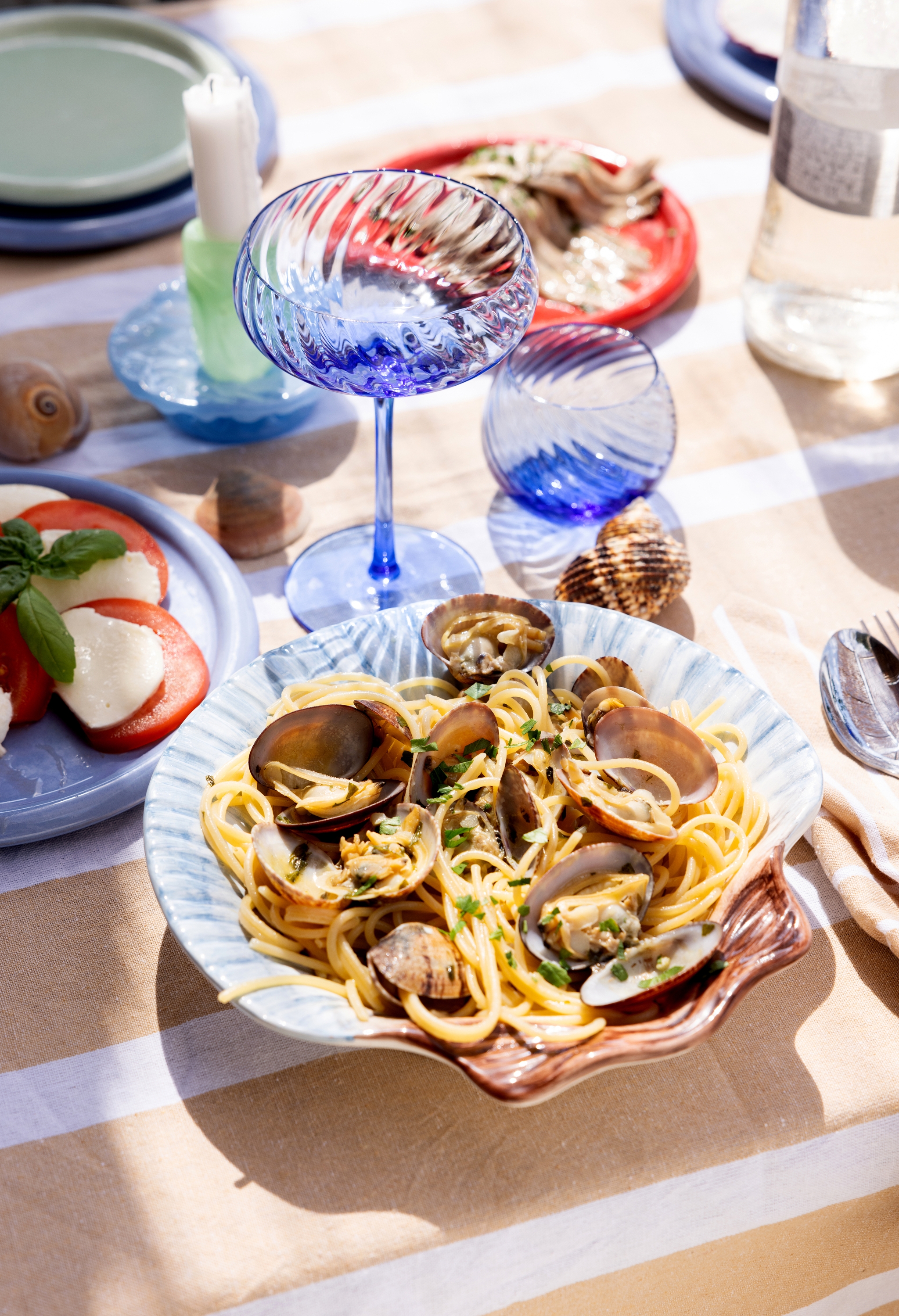 Somrig dukning med tallrik spaghetti vongole, caprese-sallad och blått glas på solbelyst bord.