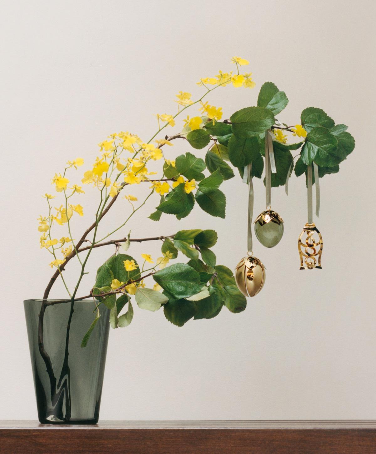 En mørkgrå vase holder grener med gule blomster og grønne blader, hvor tre dekorative påskeegg henger