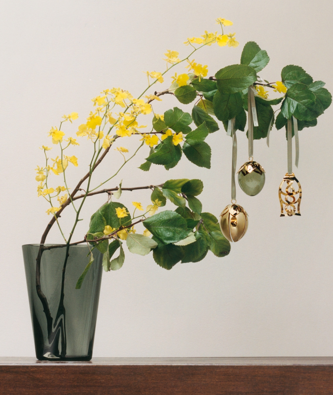 En mørkgrå vase holder grener med gule blomster og grønne blader, hvor tre dekorative påskeegg henger