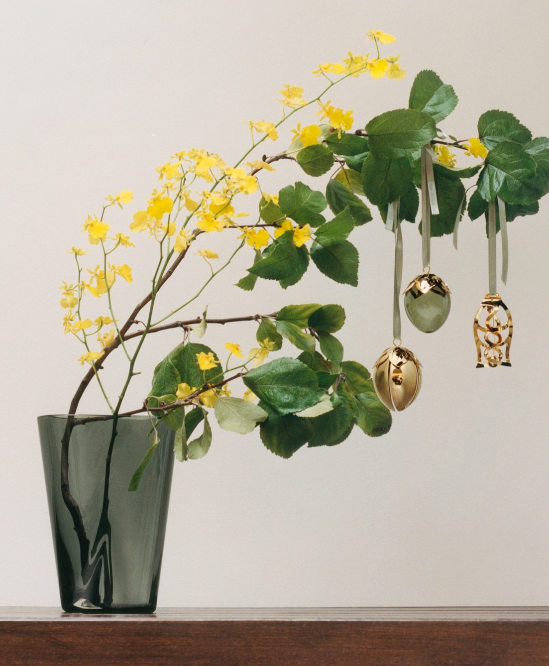 Green leaves and yellow flowers on a branch with hanging decorative ornaments, including a green egg with gold trim and a gold spiral design.