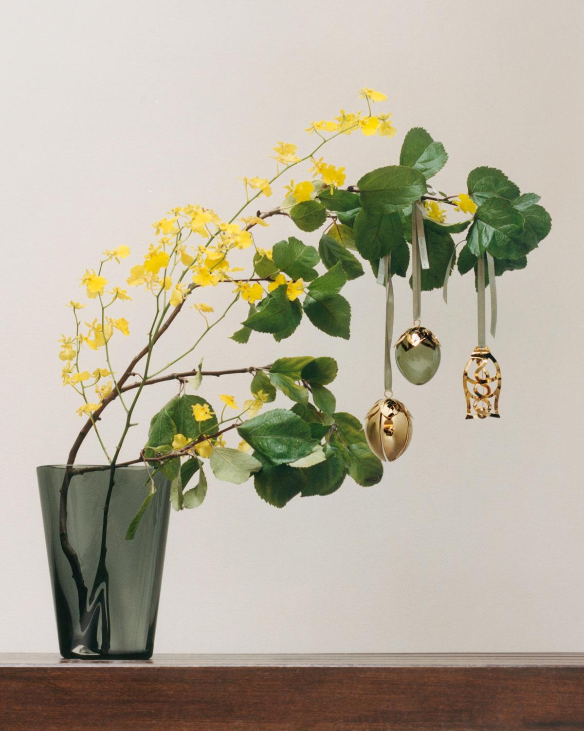 Green leaves and small yellow flowers on branches, with hanging golden bell and green glass egg ornaments.