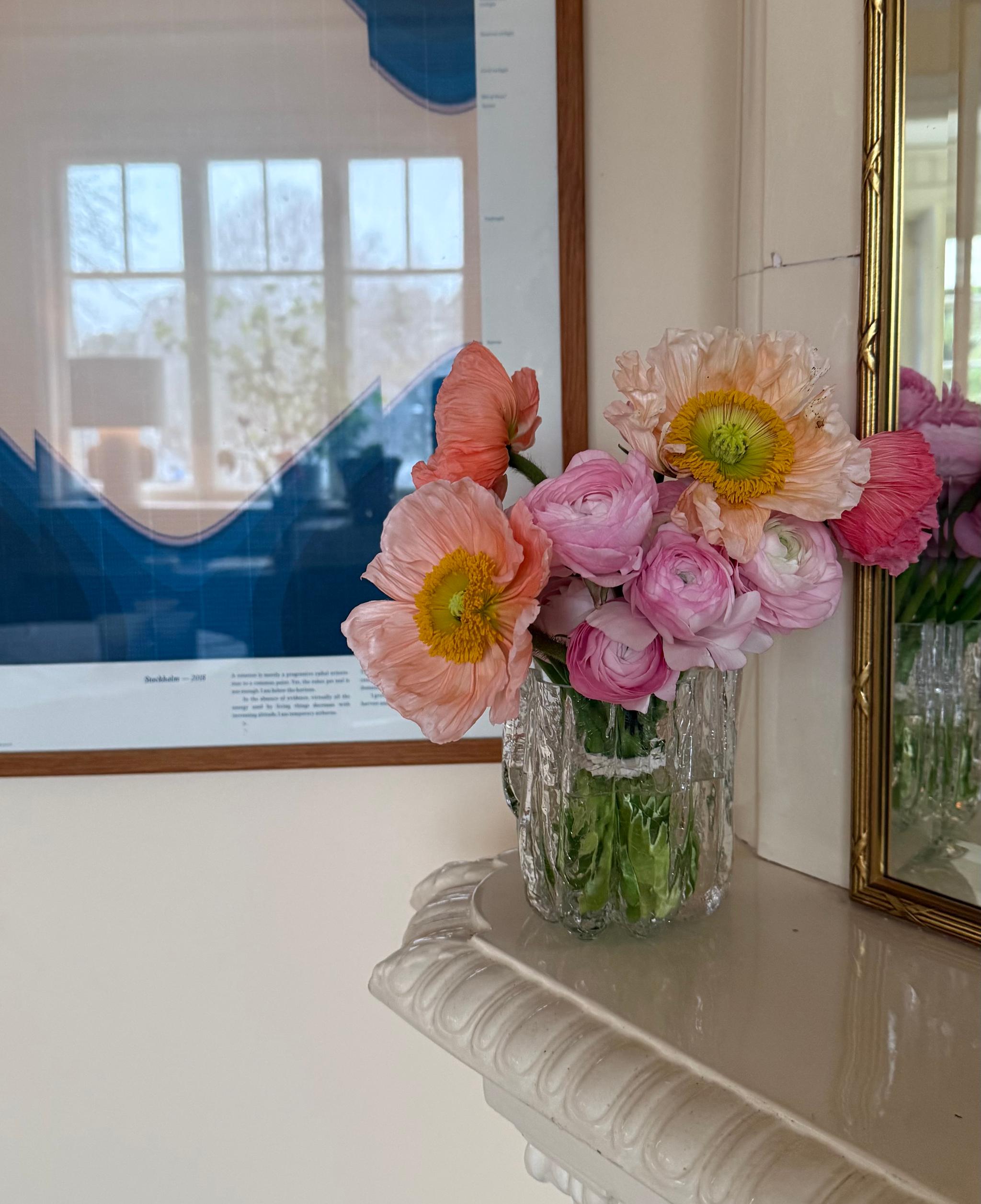 A vase of pink and orange flowers sits on a white mantelpiece, with a blue abstract art print and a golden mirror in the background.