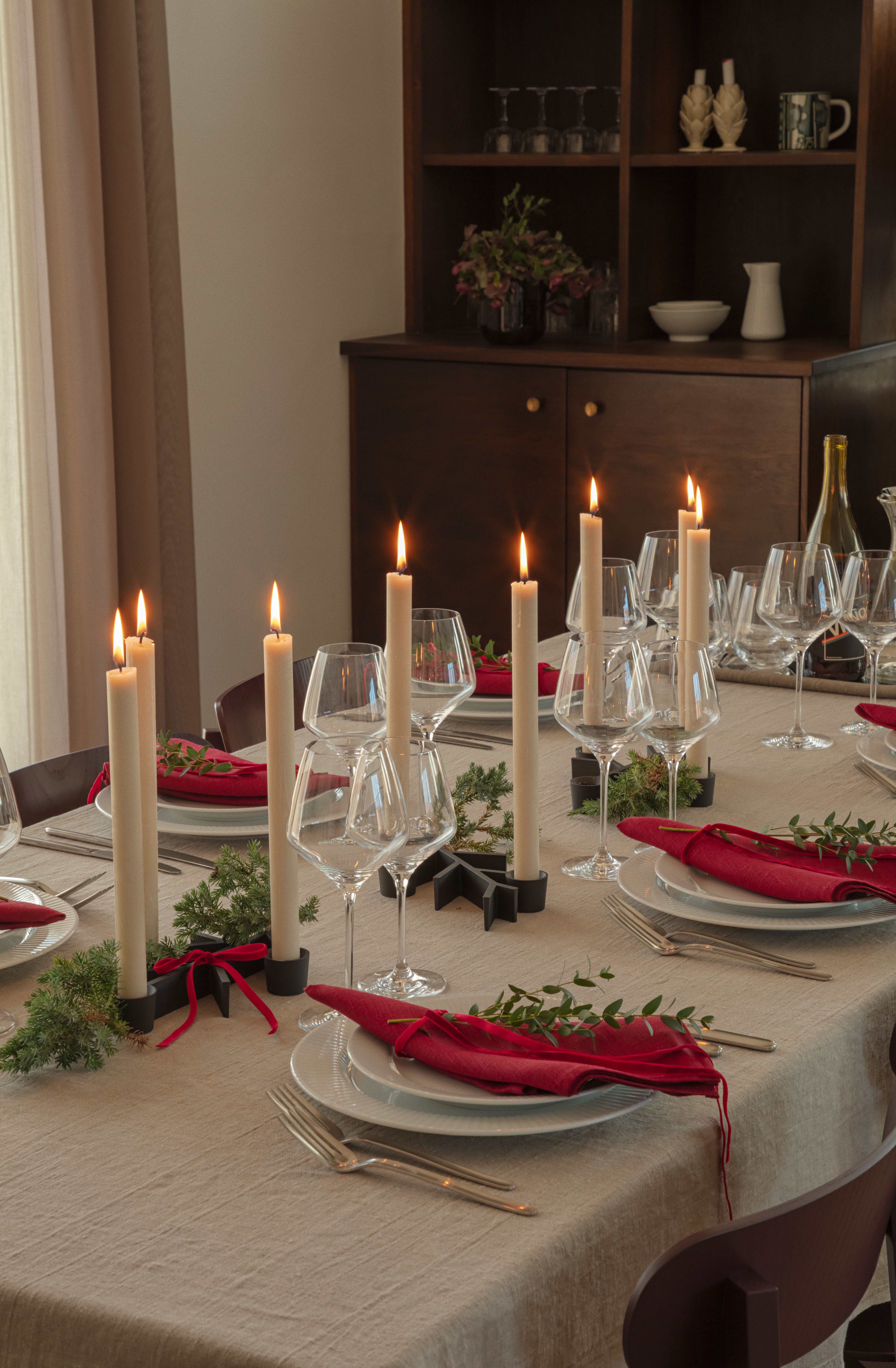 Juhlava ruokapöytä, jossa on kynttilöitä, punaisia ​​lautasliinoja, viinilaseja ja ikivihreitä kasveja. Taustalla tumma kaappi astioineen.