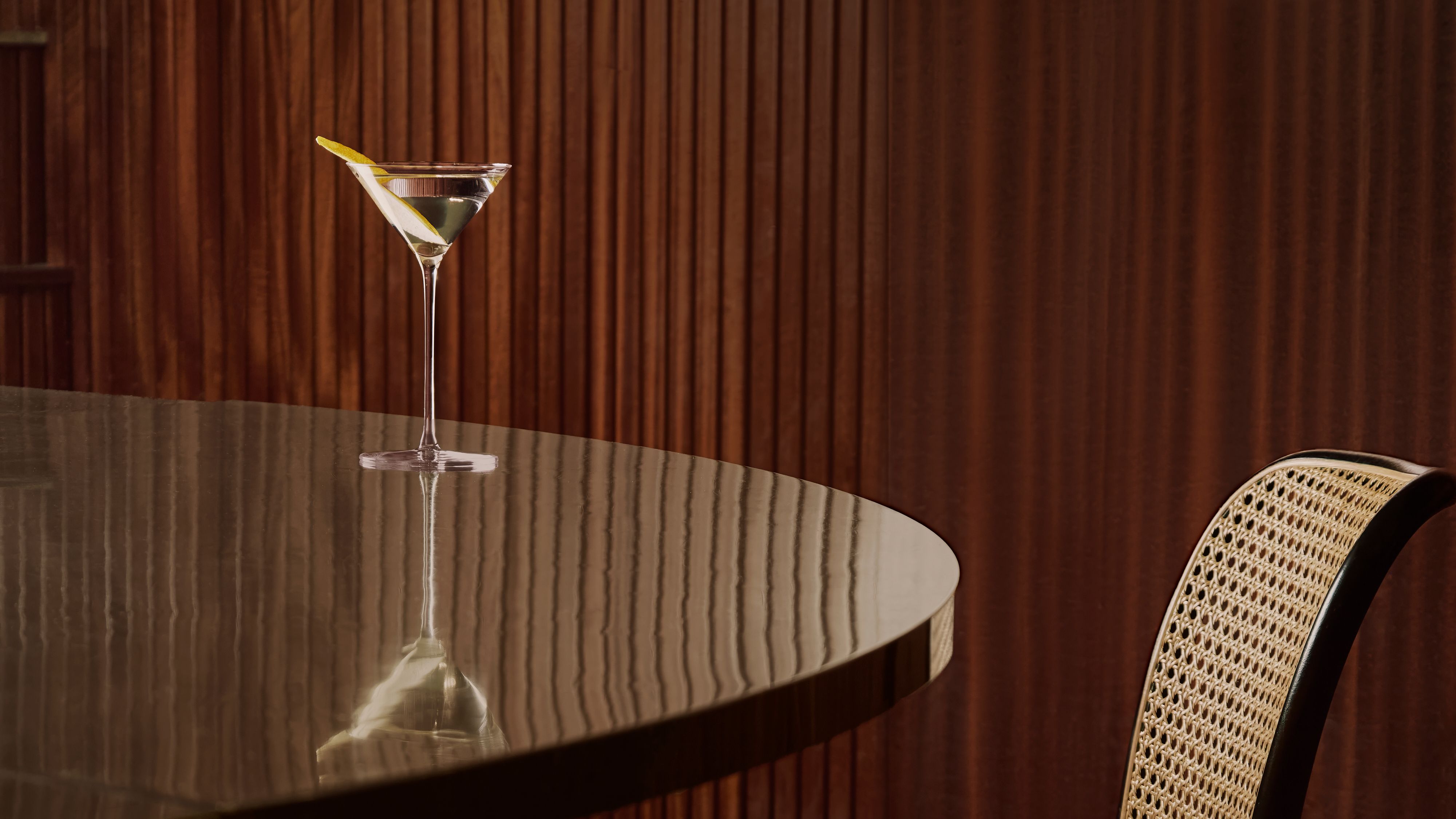 Martini med en sitrontwist på et reflekterende bord ved siden av en trepanelvegg.