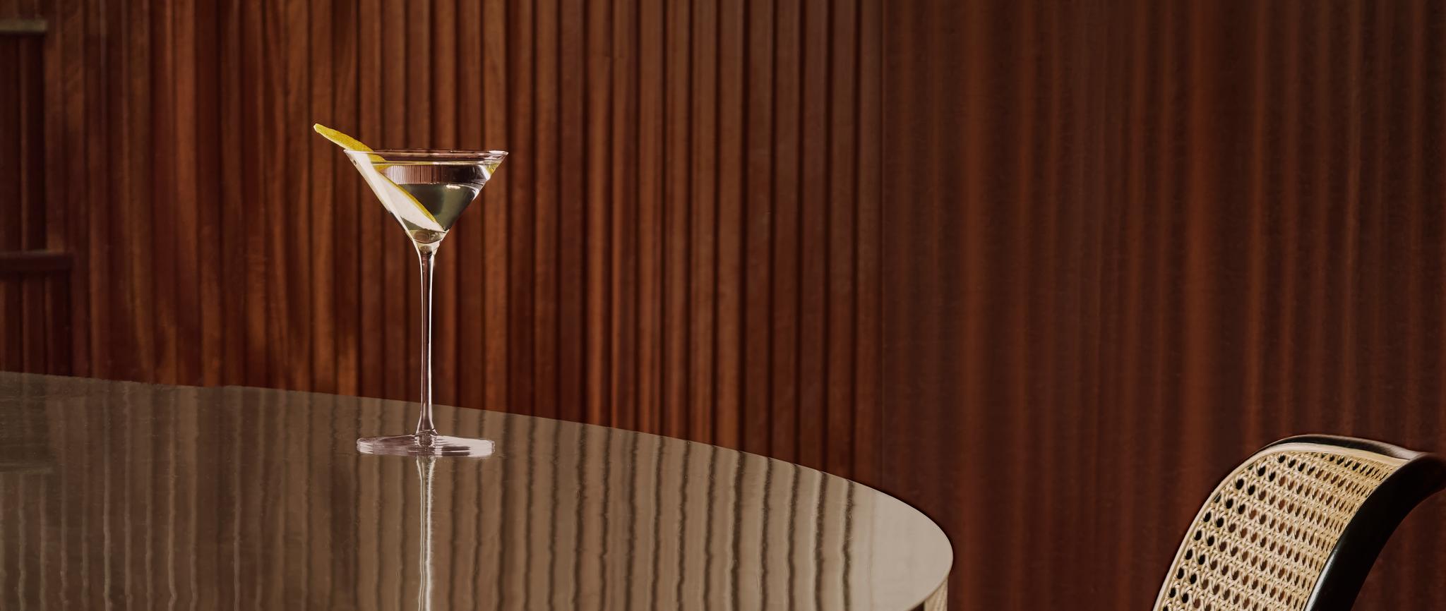 Sitruunaisella kierteellä varustettu martinilasi heijastavalla pöydällä rottinkituolin ja puupaneeliseinän vieressä.
