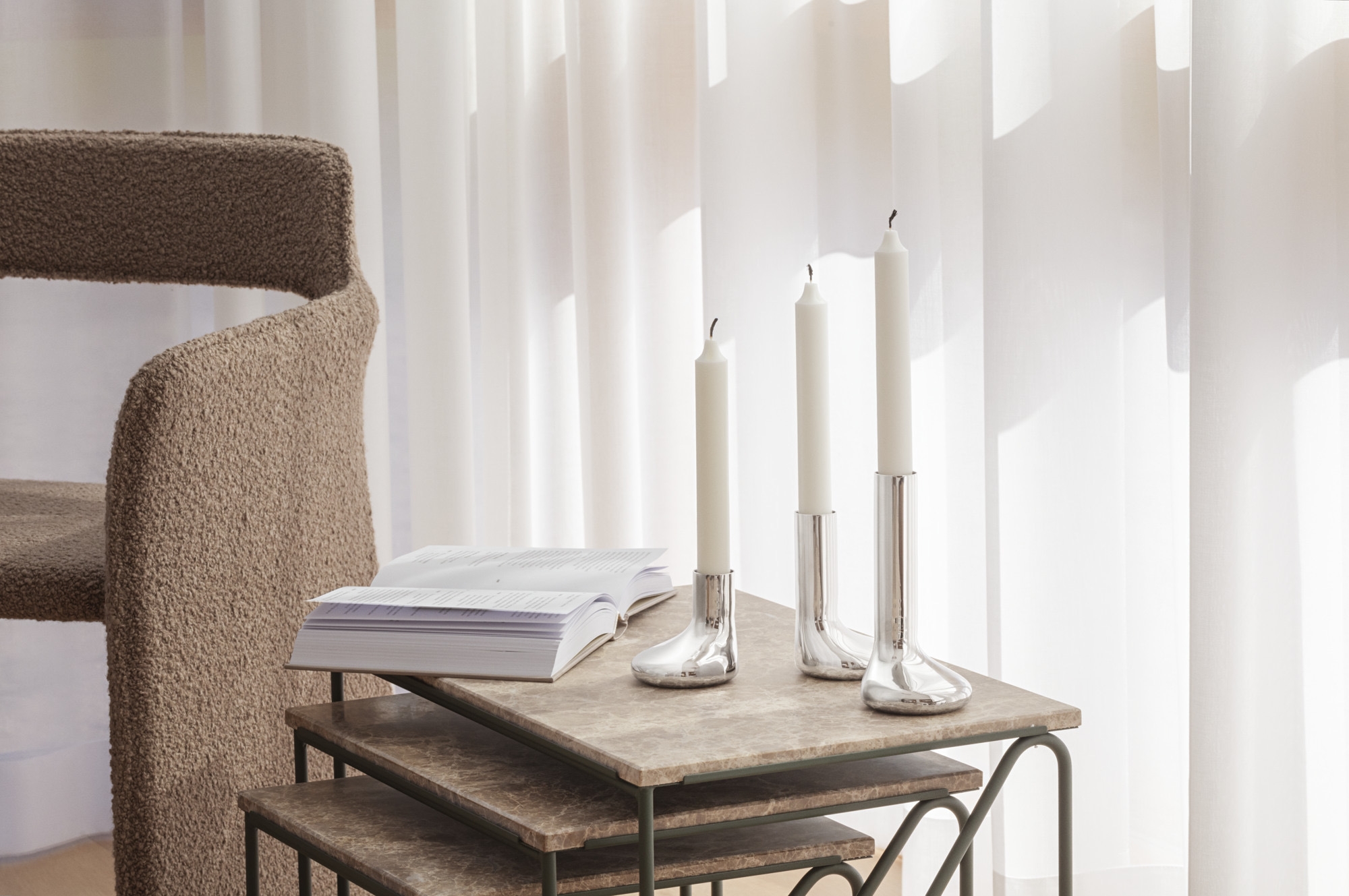 Three white candles in silver holders, an open book, and a textured brown chair on a nesting table.