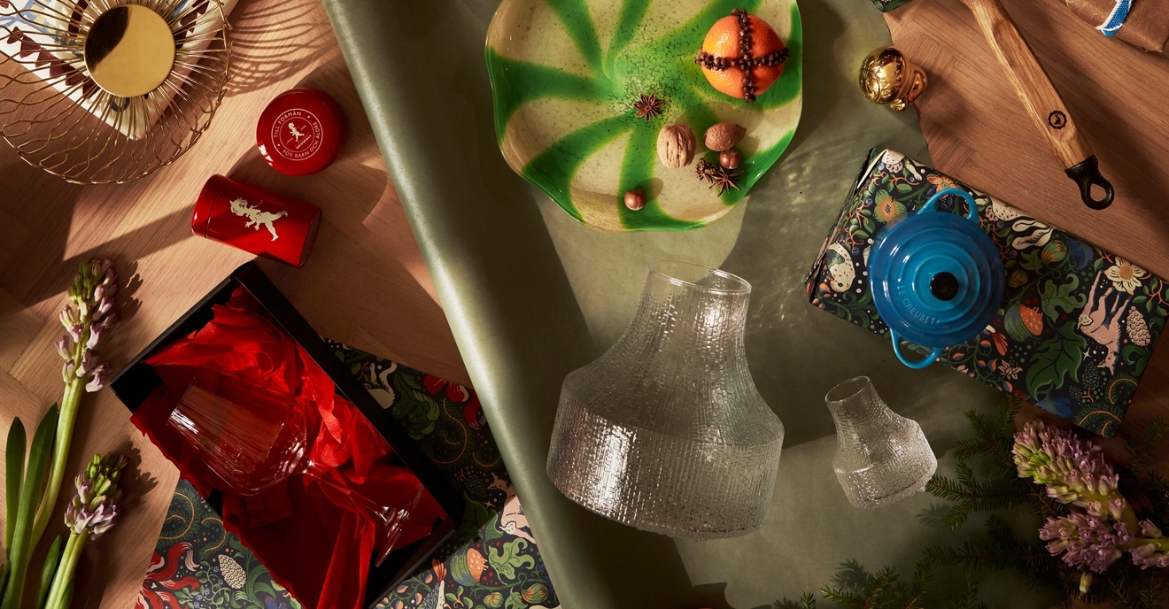 Overhead flat lay of diverse holiday gifts and kitchen items, including a knife set, cast iron skillet, and glassware, on patterned paper and wood.