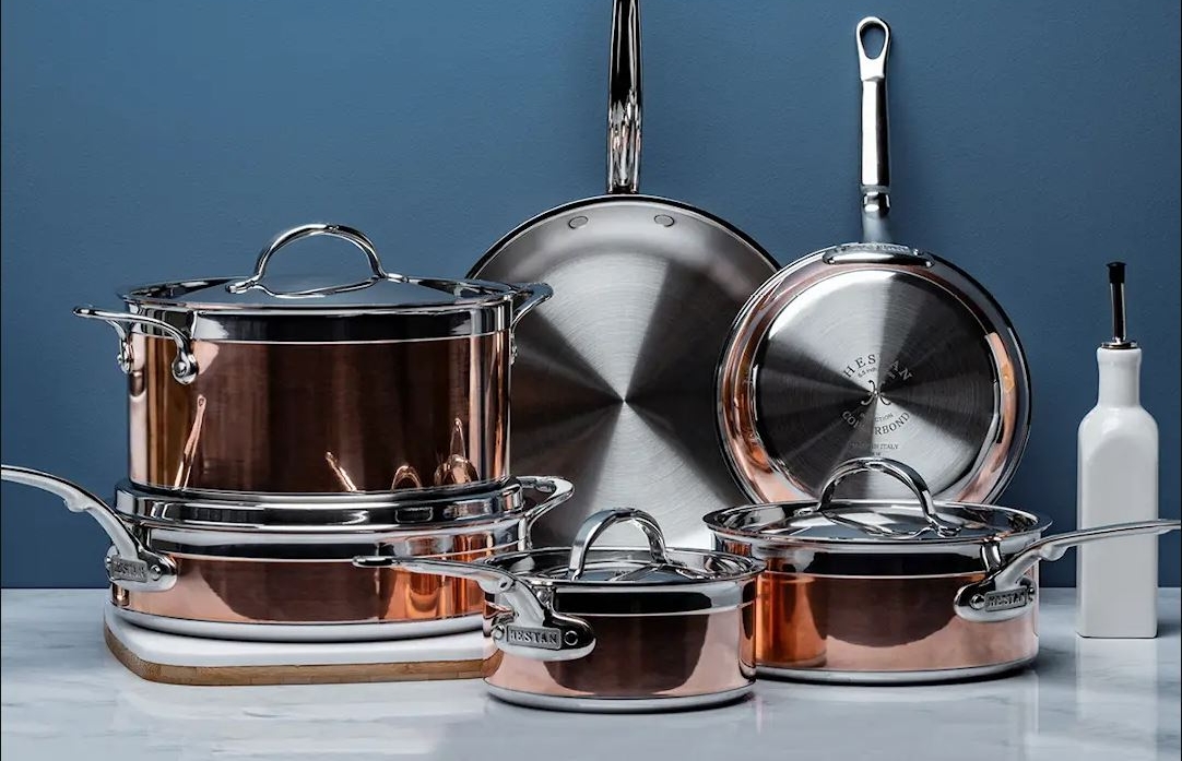 A set of shiny copper and stainless steel cookware on a marble surface against a blue wall.
