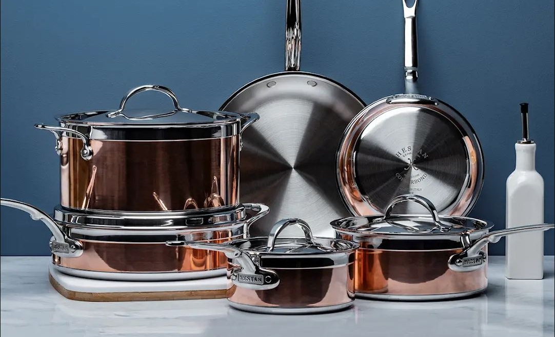 A collection of copper and stainless steel pots and pans, with a white oil dispenser, on a marble surface against a blue wall.