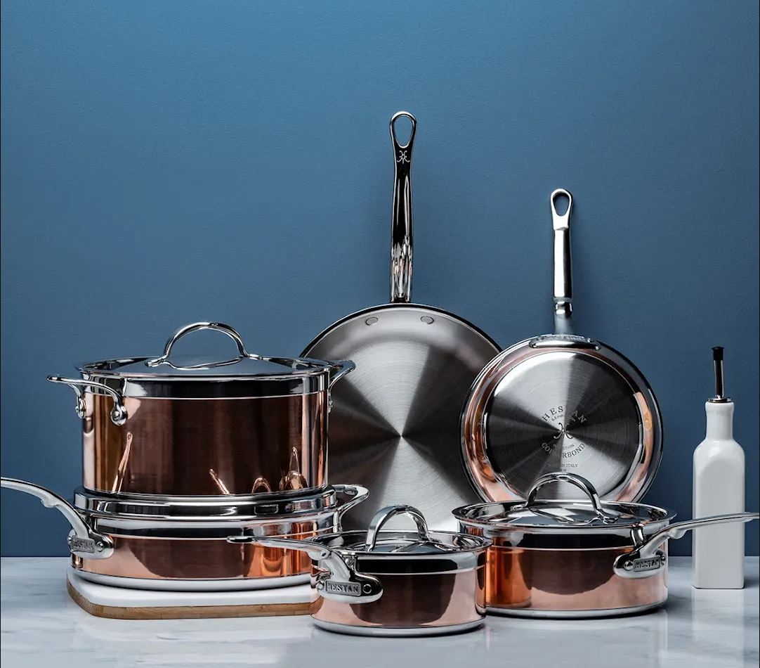 A set of shiny copper and stainless steel cookware on a marble surface against a blue wall.