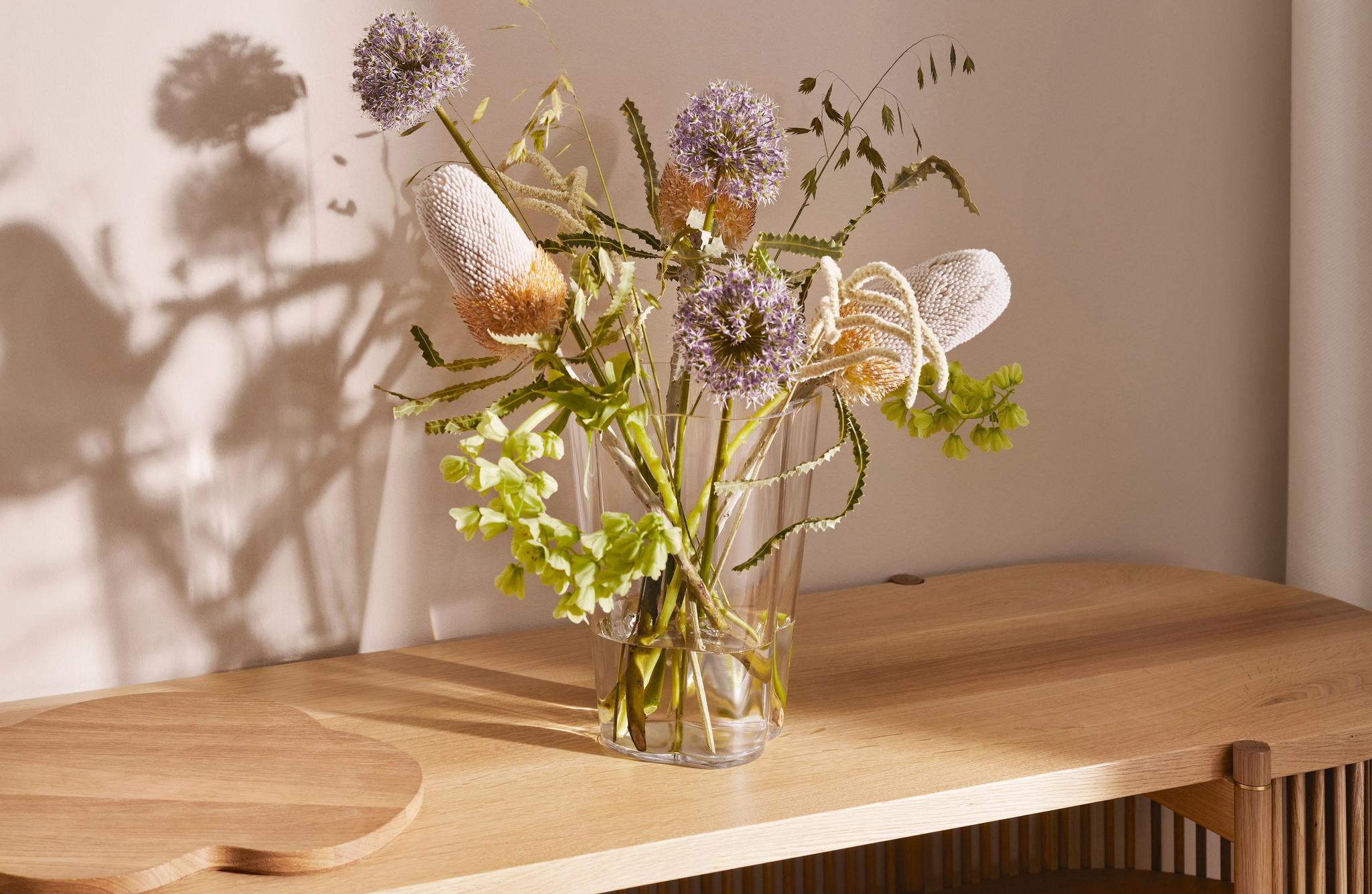 Vaalealla puisella konsolipöydällä on maljakko, jossa on violetteja ja kermanvärisiä kukkia, ja sen takana olevalle seinälle heittyy kasvien varjoja.