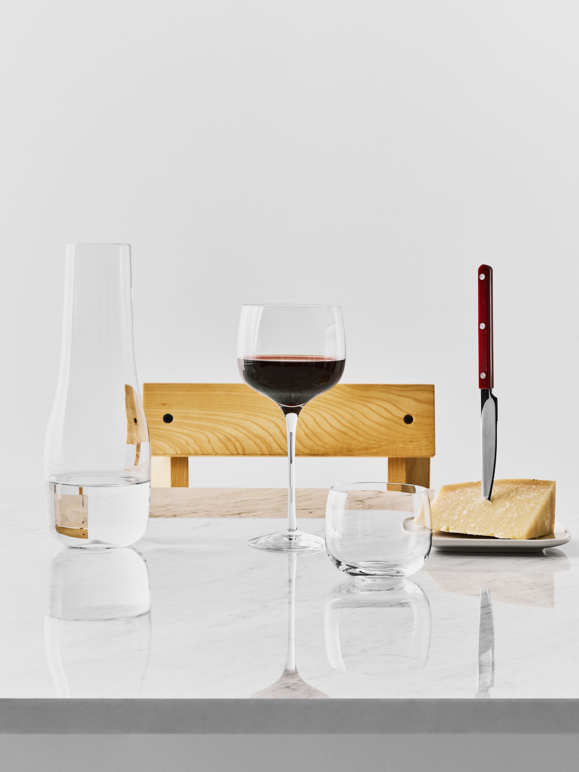 Stilleben med ett glas rött vin, vattenkaraff och parmesanost med kniv på ett ljust marmorbord.