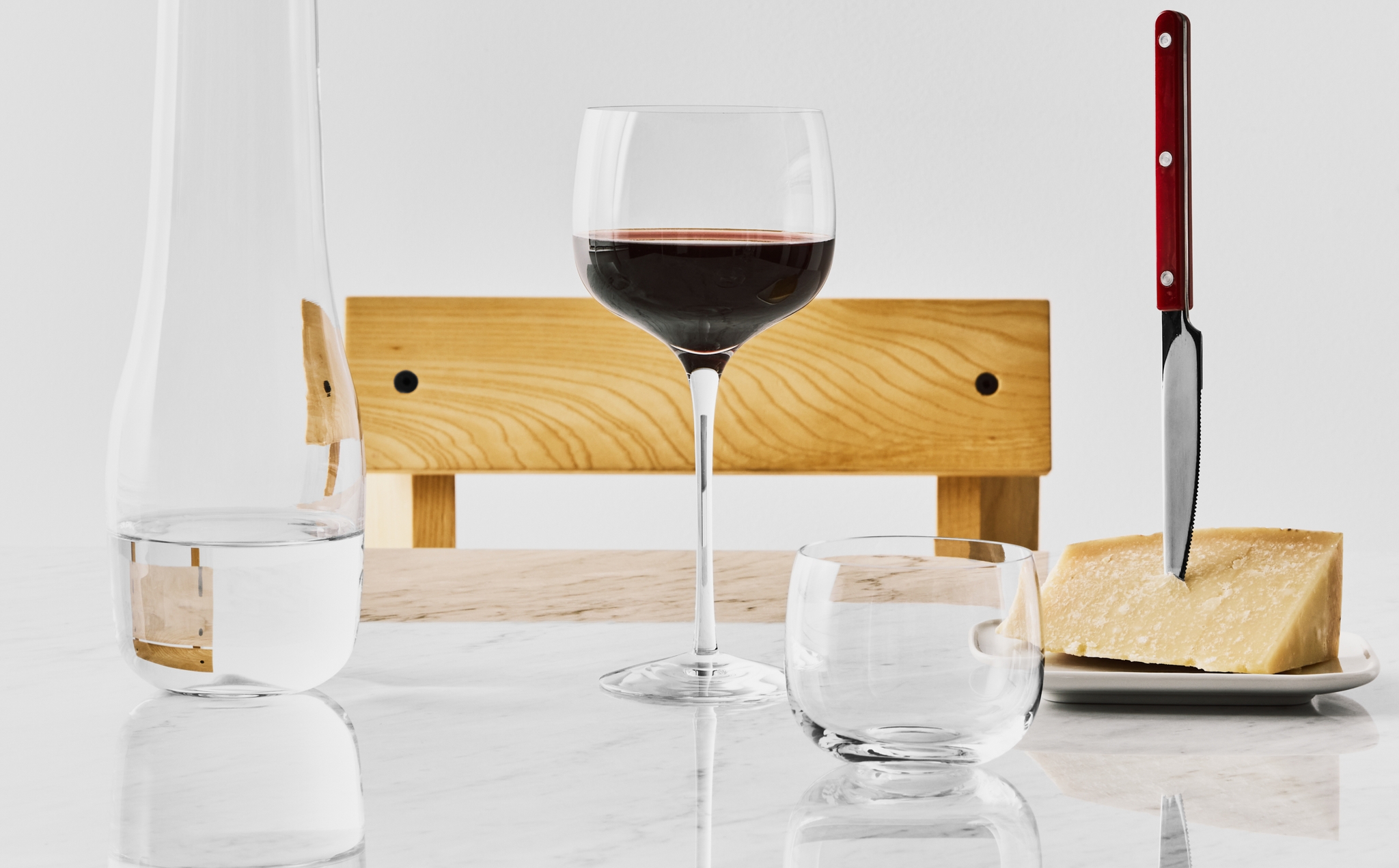 Stilleben med ett glas rött vin, vattenkaraff och parmesanost med kniv på ett ljust marmorbord.