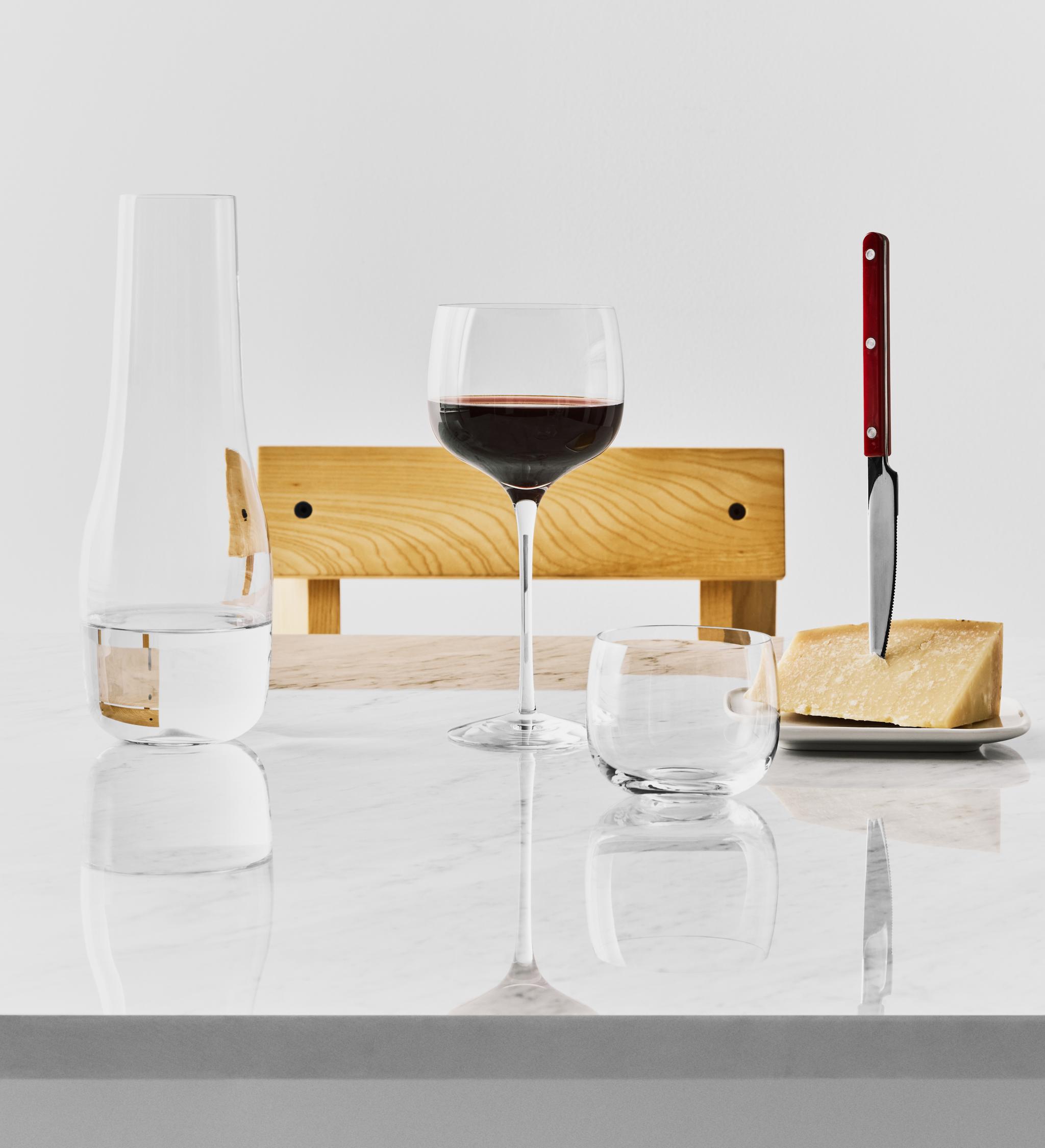 Stilleben med ett glas rött vin, vattenkaraff och parmesanost med kniv på ett ljust marmorbord.