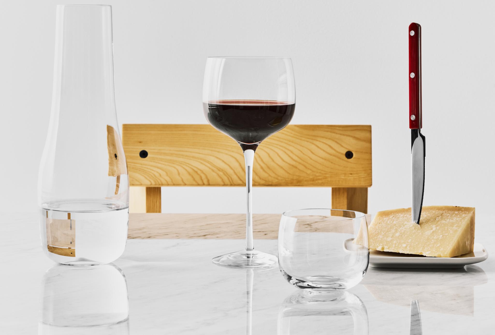 Stilleben med ett glas rött vin, vattenkaraff och parmesanost med kniv på ett ljust marmorbord.