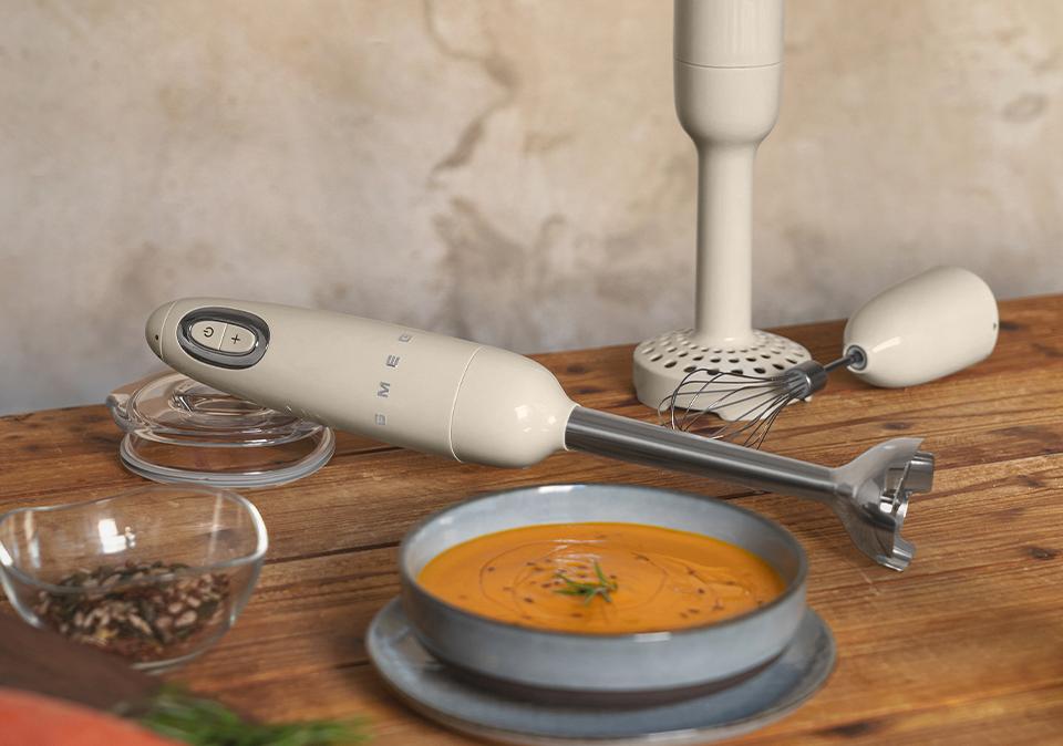 En cremefärgad stavmixer från Smeg ligger på ett träbord bredvid en tallrik med soppa