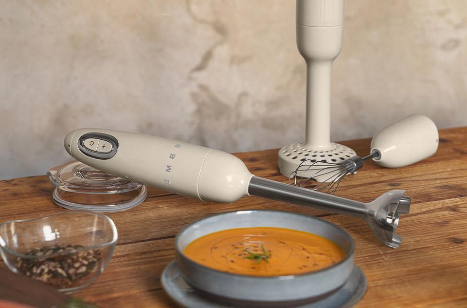 Kermanvärinen Smeg-sauvansekoitin, vispiläosa ja kulho appelsiinikeittoa puupöydällä.