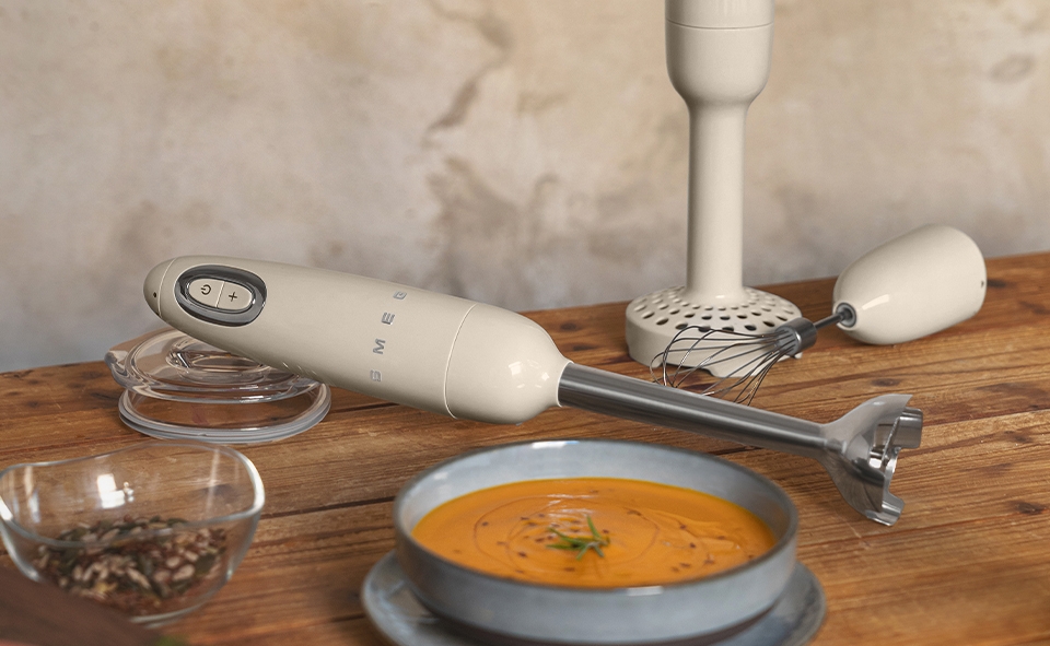 En Smeg-stavmixer och visptillbehör står på ett träbord med en skål morotssoppa, råa morötter och kryddor.