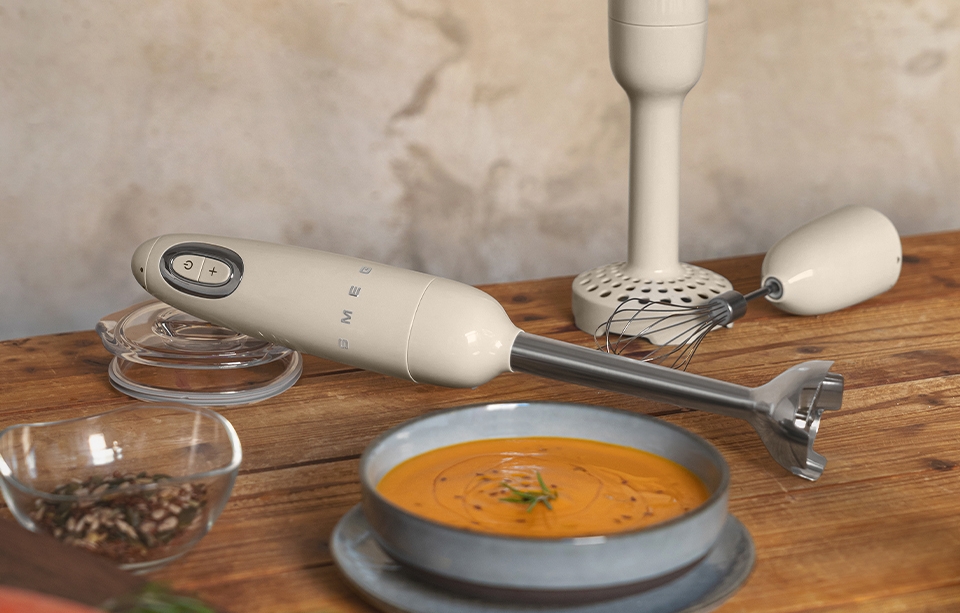 En Smeg-stavmixer och visptillbehör står på ett träbord med en skål morotssoppa, råa morötter och kryddor.