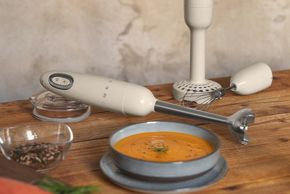 Smeg-sauvasekoitin ja vispiläosa ovat puupöydällä kulhollisen porkkanakeittoa, raakoja porkkanoita ja mausteita kanssa.