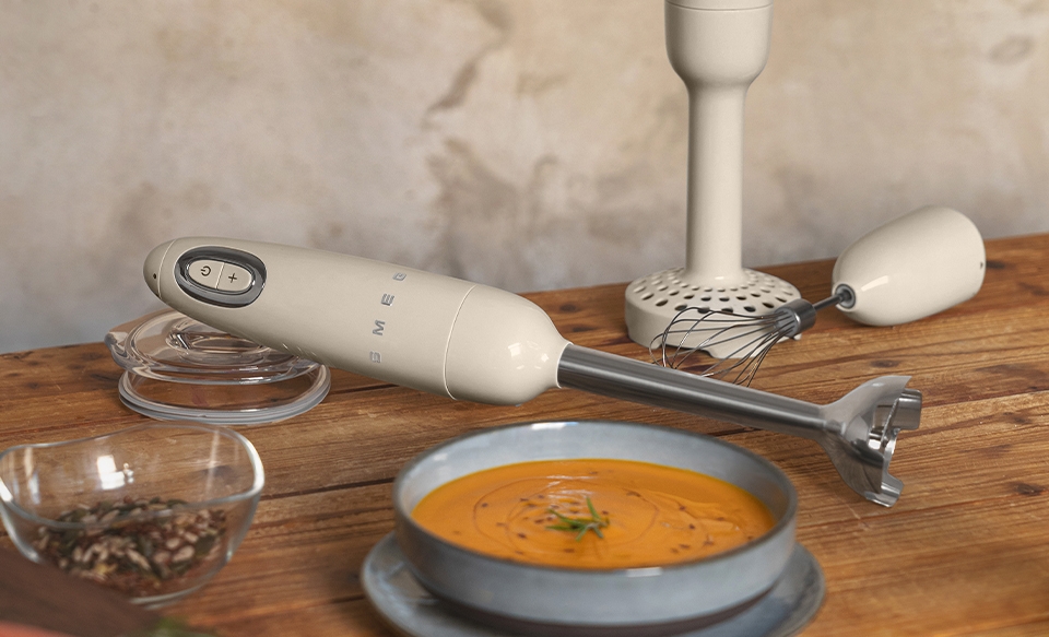 Smeg-sauvasekoitin ja vispiläosa ovat puupöydällä kulhollisen porkkanakeittoa, raakoja porkkanoita ja mausteita kanssa.