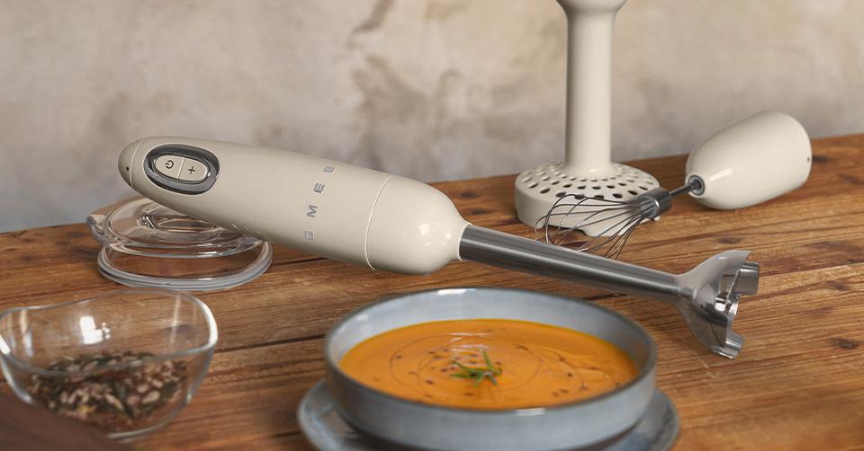 Sauvasekoitin ja sen lisäosat puupöydällä kirkkaan oranssin keittokulhon vieressä.