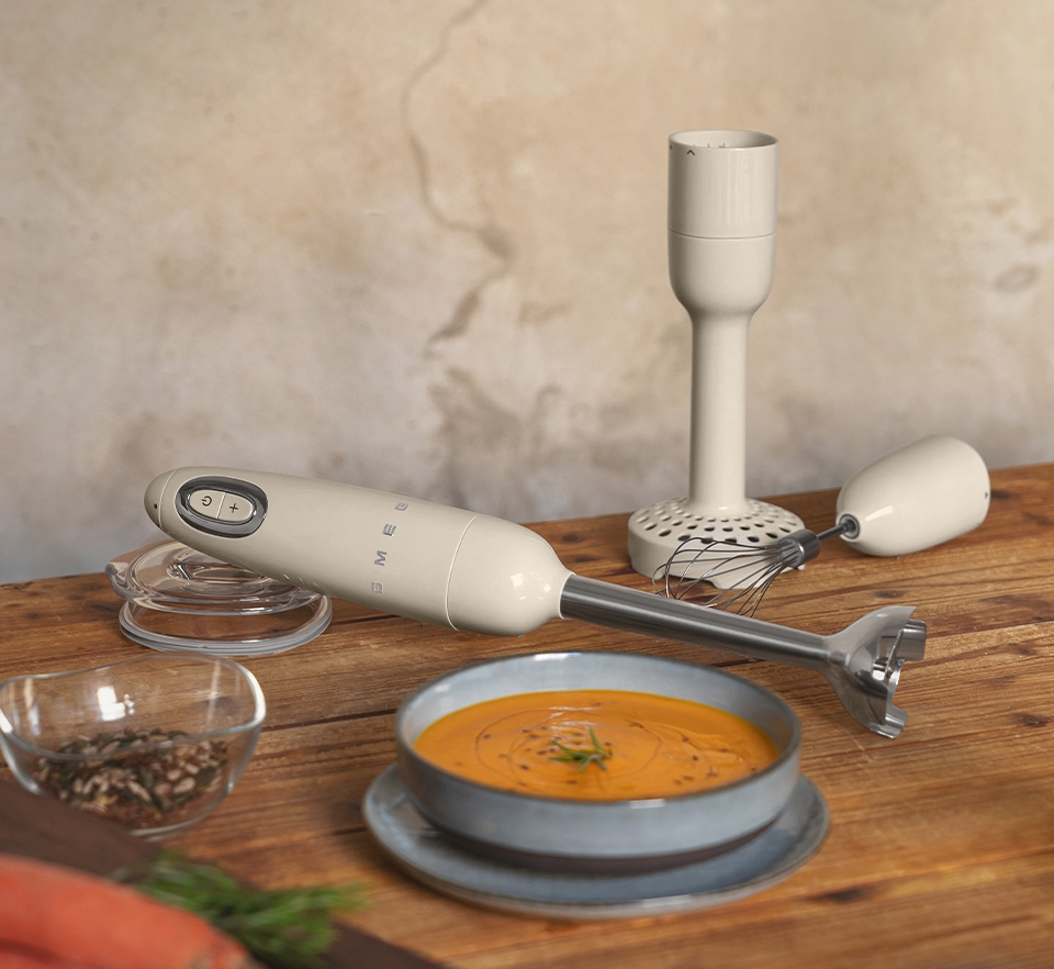 En cremefärgad stavmixer från Smeg ligger på ett träbord bredvid en tallrik med soppa