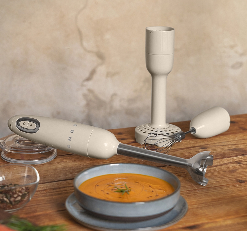 Sauvasekoitin ja sen lisäosat puupöydällä kirkkaan oranssin keittokulhon vieressä.