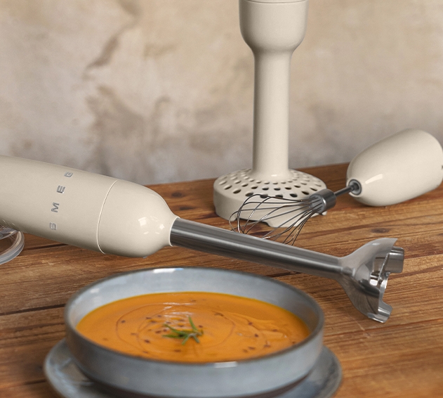 Kermanvärinen Smeg-sauvansekoitin, vispiläosa ja kulho appelsiinikeittoa puupöydällä.