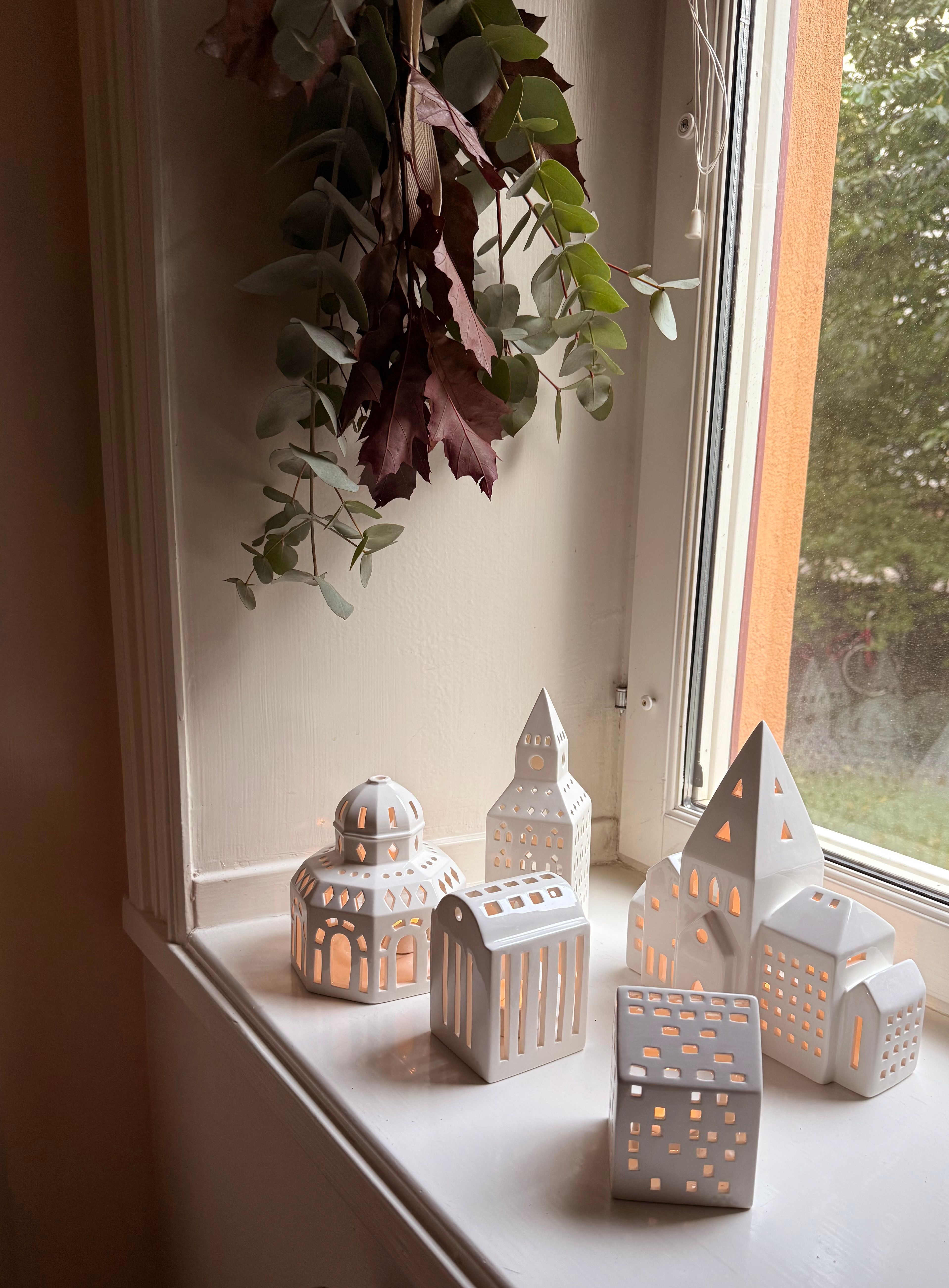 White ceramic miniature buildings and lanterns with glowing lights on a windowsill, beneath dried eucalyptus and red leaves.
