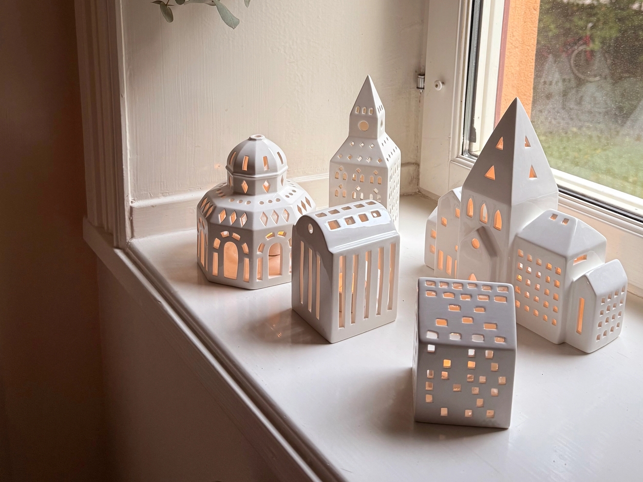 White ceramic house and lantern-shaped candle holders with lit candles on a window sill, below hanging leaves.