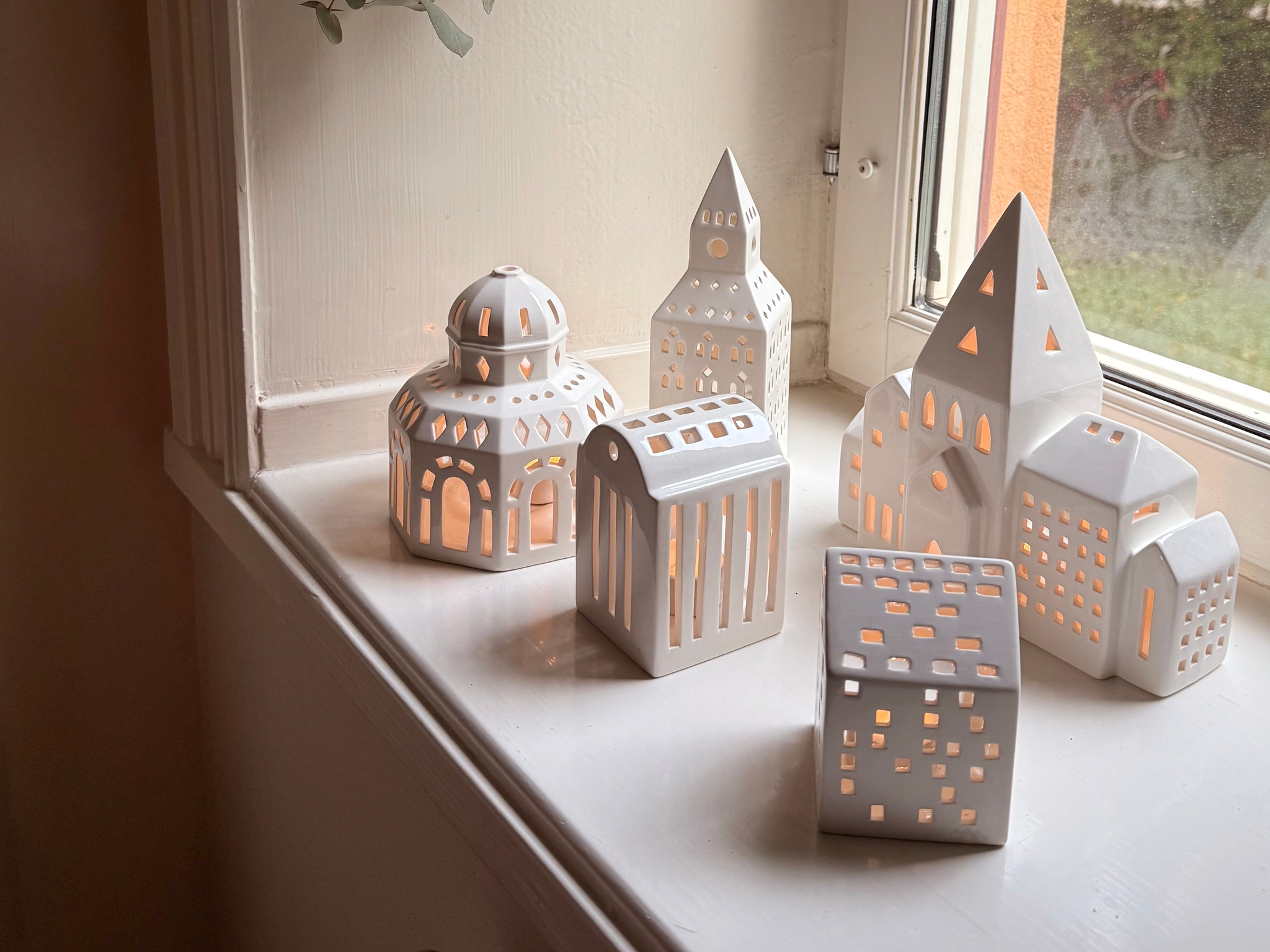 White ceramic house and lantern-shaped candle holders with lit candles on a window sill, below hanging leaves.