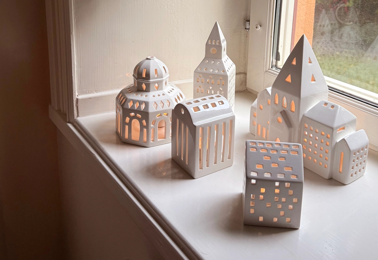 White ceramic miniature buildings and lanterns with glowing lights on a windowsill, beneath dried eucalyptus and red leaves.